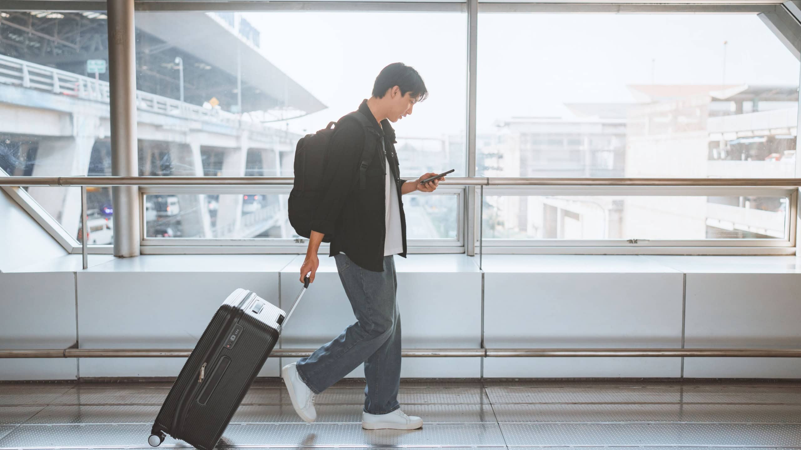 Side view - Male traveler walking with luggage using mobile smartphone at the modern airport terminal, man on way to flight boarding gate, Ready for travel or vacation journey, copy space