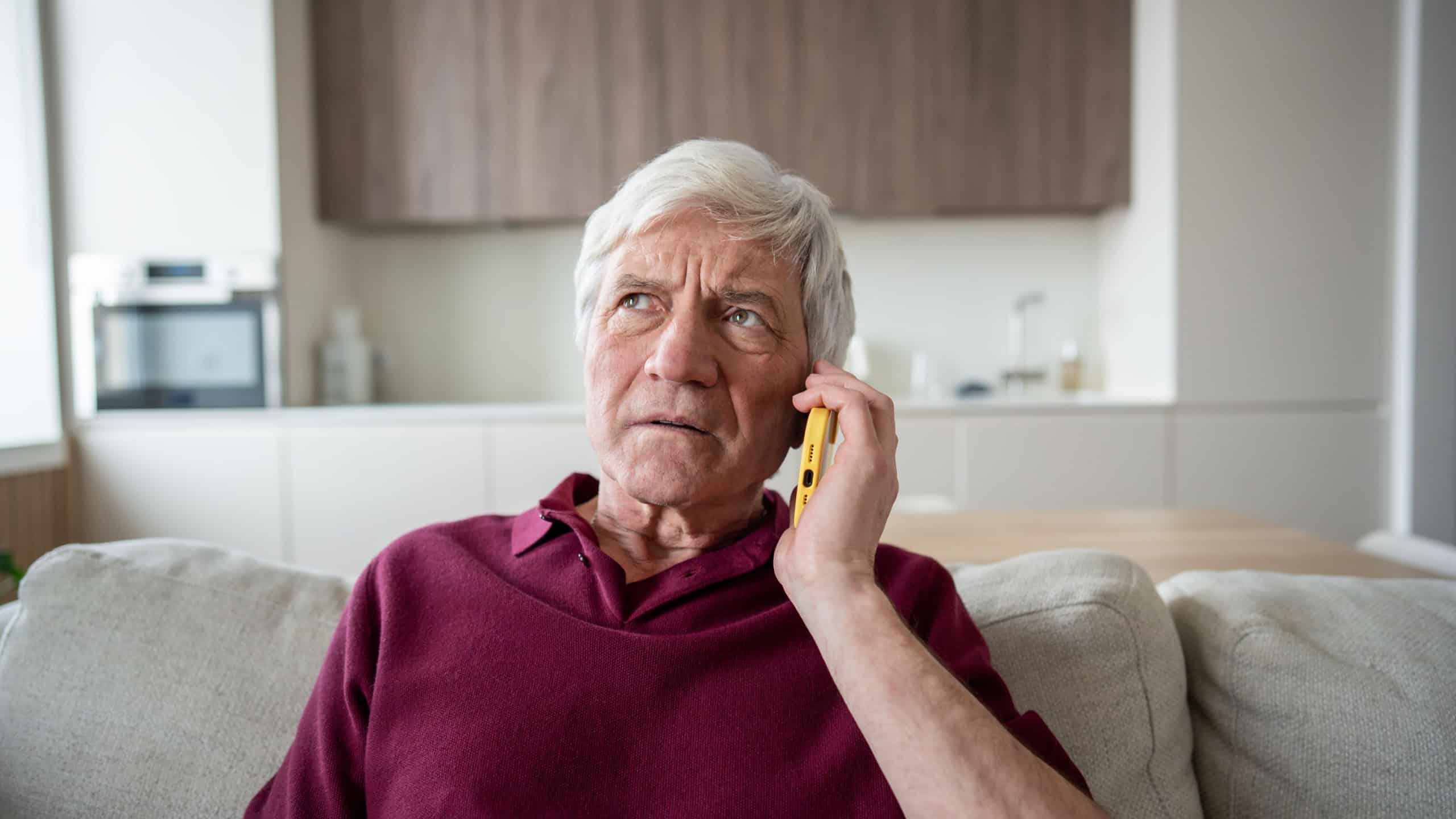 Distrustful and anxious senior man frowns during emergency call, listening to pressured demands for banking information, sitting at sofa. Fraud warning, mislead, personal data risk, financial scam