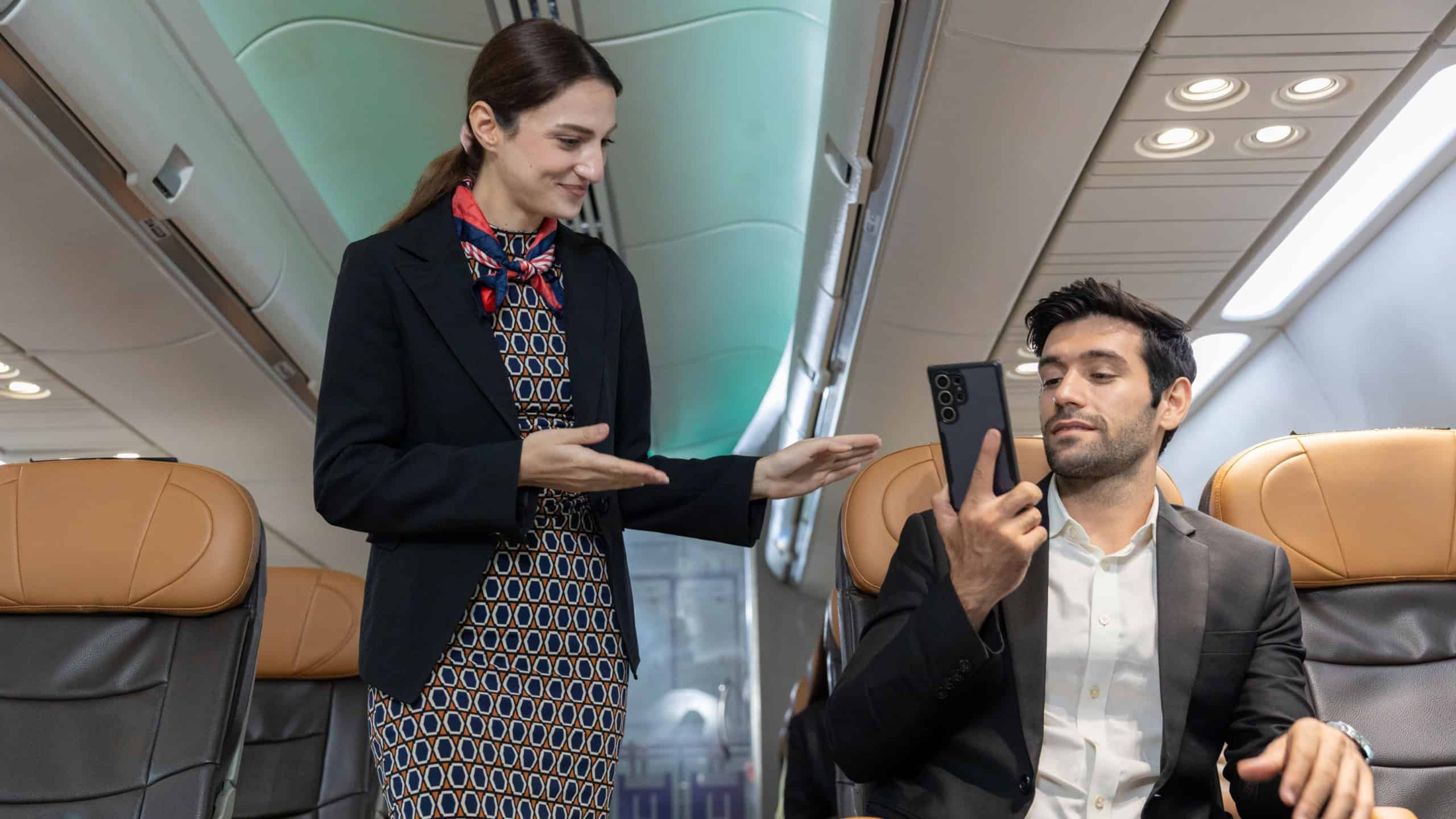 Flight attendant interacts with male passenger talking on mobile phone during boarding or pre-flight procedure inside commercial airplane cabin.