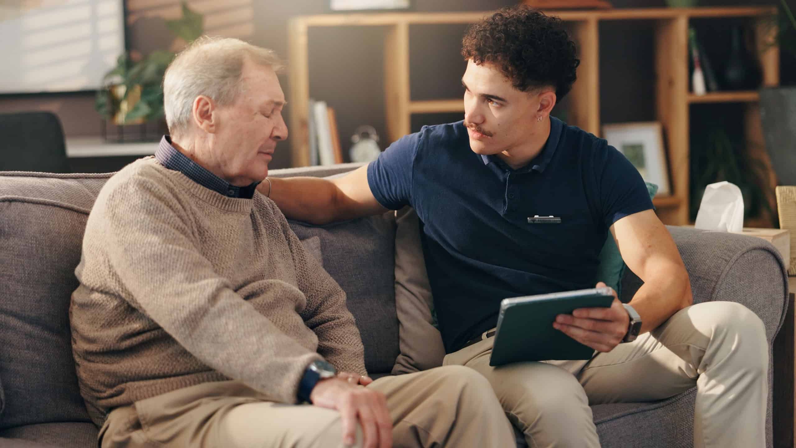 Old man, nurse and tablet on sofa, sad and report with diagnosis in living room in home. Caregiver, elderly person and digital touchscreen with comfort, app and results with bad news in retirement