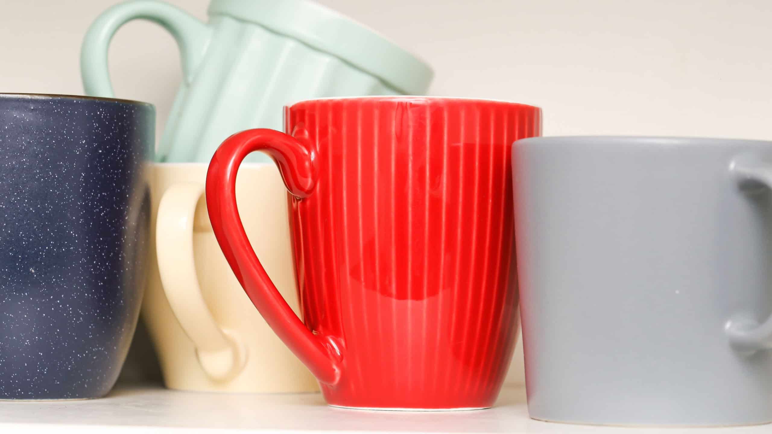 Colorful ceramic mugs on white shelf. Assorted kitchen cups in different shapes and colors. Red, blue, gray, mint and beige mugs. Stack of coffee cups. Interior detail from home kitchen decor.