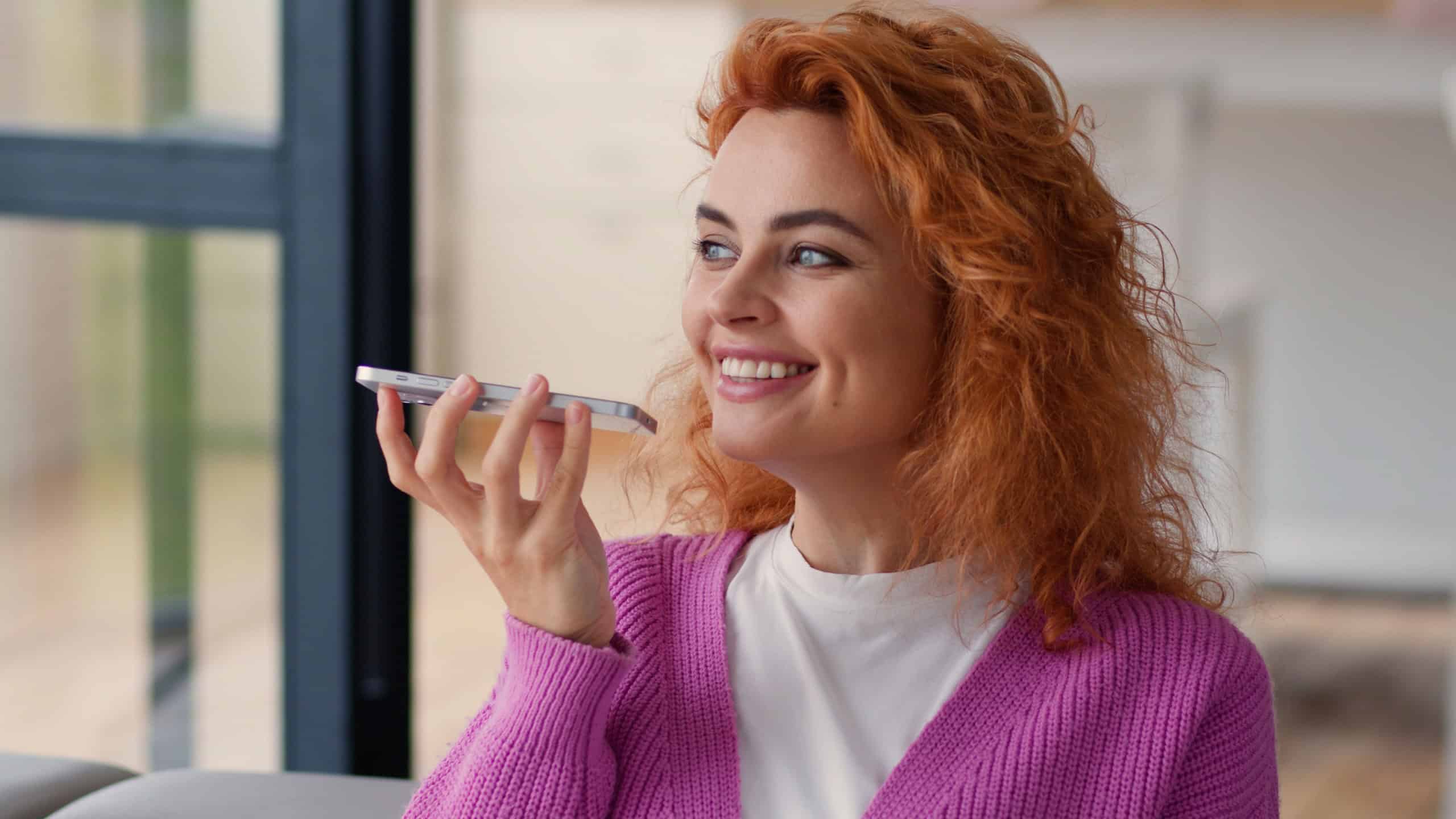 Happy smiling carefree Caucasian woman lady girl indoors at home couch sofa female using mobile phone app network smile record audio message recording voice call message talk to microphone smartphone