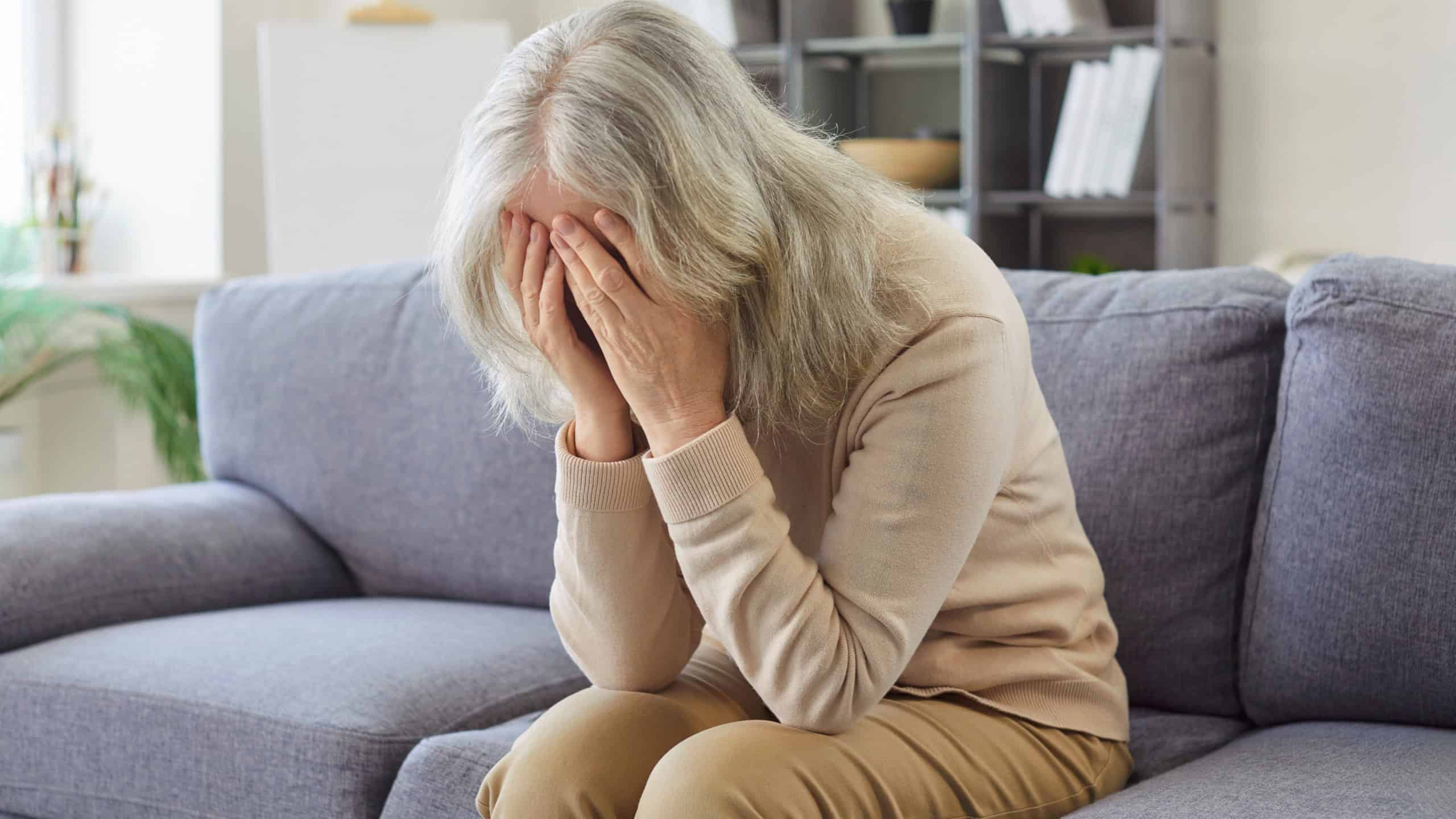 Old senior woman sitting indoors on sofa in distress at home, covering face with hands, weeping feeling overwhelmed, sadness or emotional pain, feeling lonely, lacking social or emotional connection
