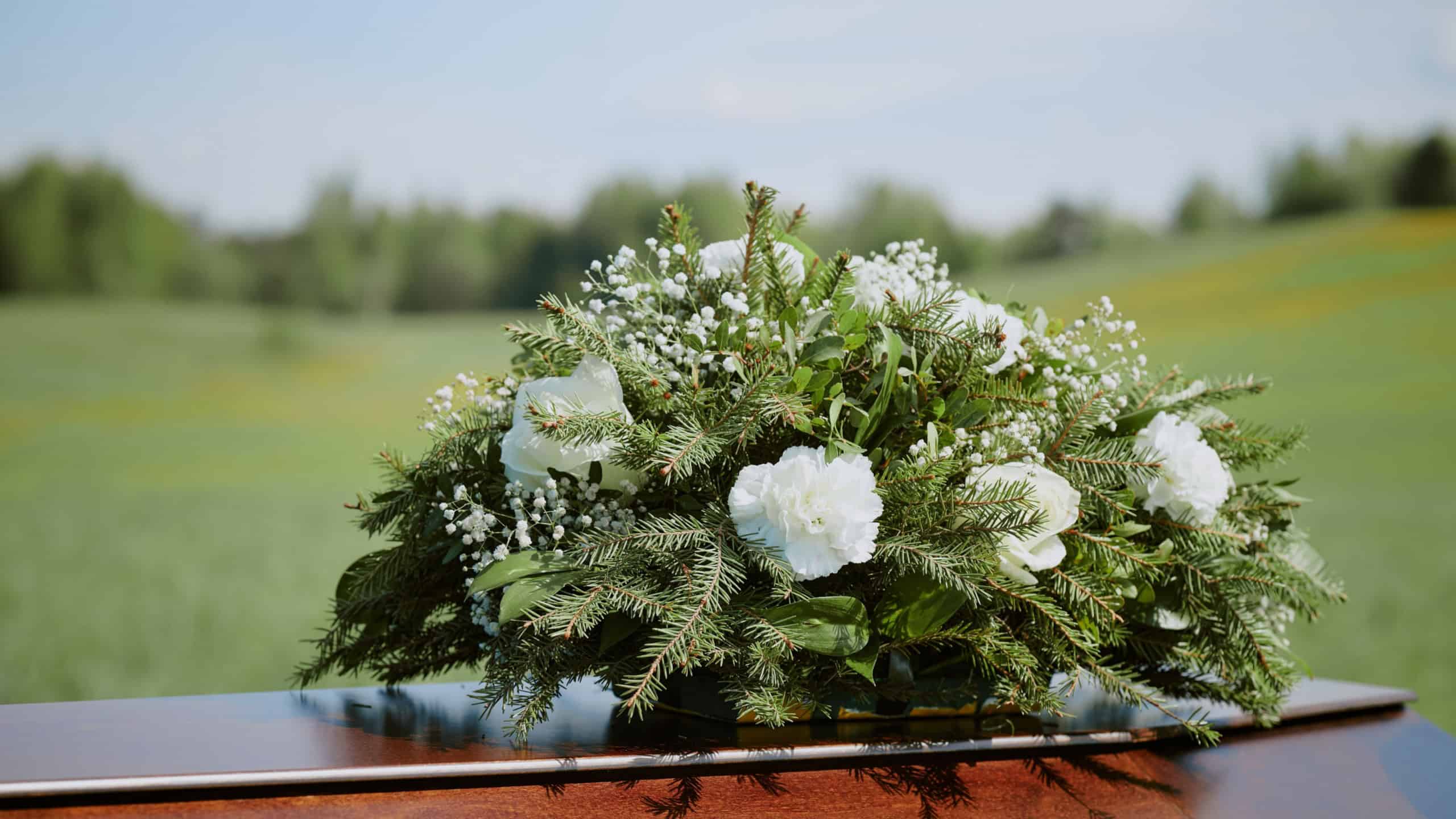 Funeral casket adorned with flowers and greenery resting outdoors on grassy field, floral arrangement featuring roses and baby's breath symbolizing remembrance and mourning