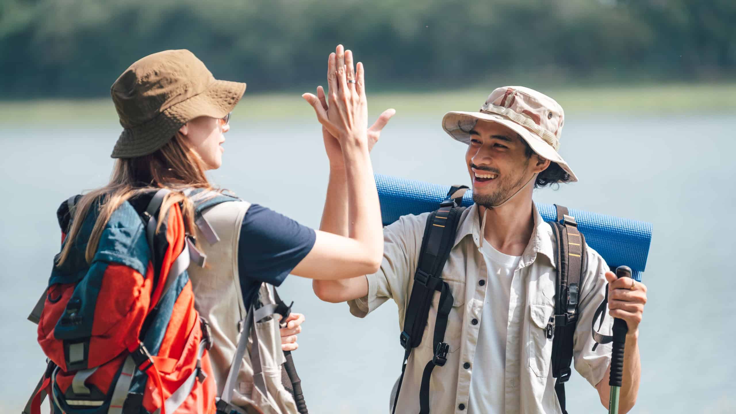 Hiking Couple Share High Five During Trekking Trip in Summer Meadow by the Lake. Man and Woman Backpackers Show Teamwork, Friendship, and Adventure Spirit in Outdoor Journey Surrounded by Nature