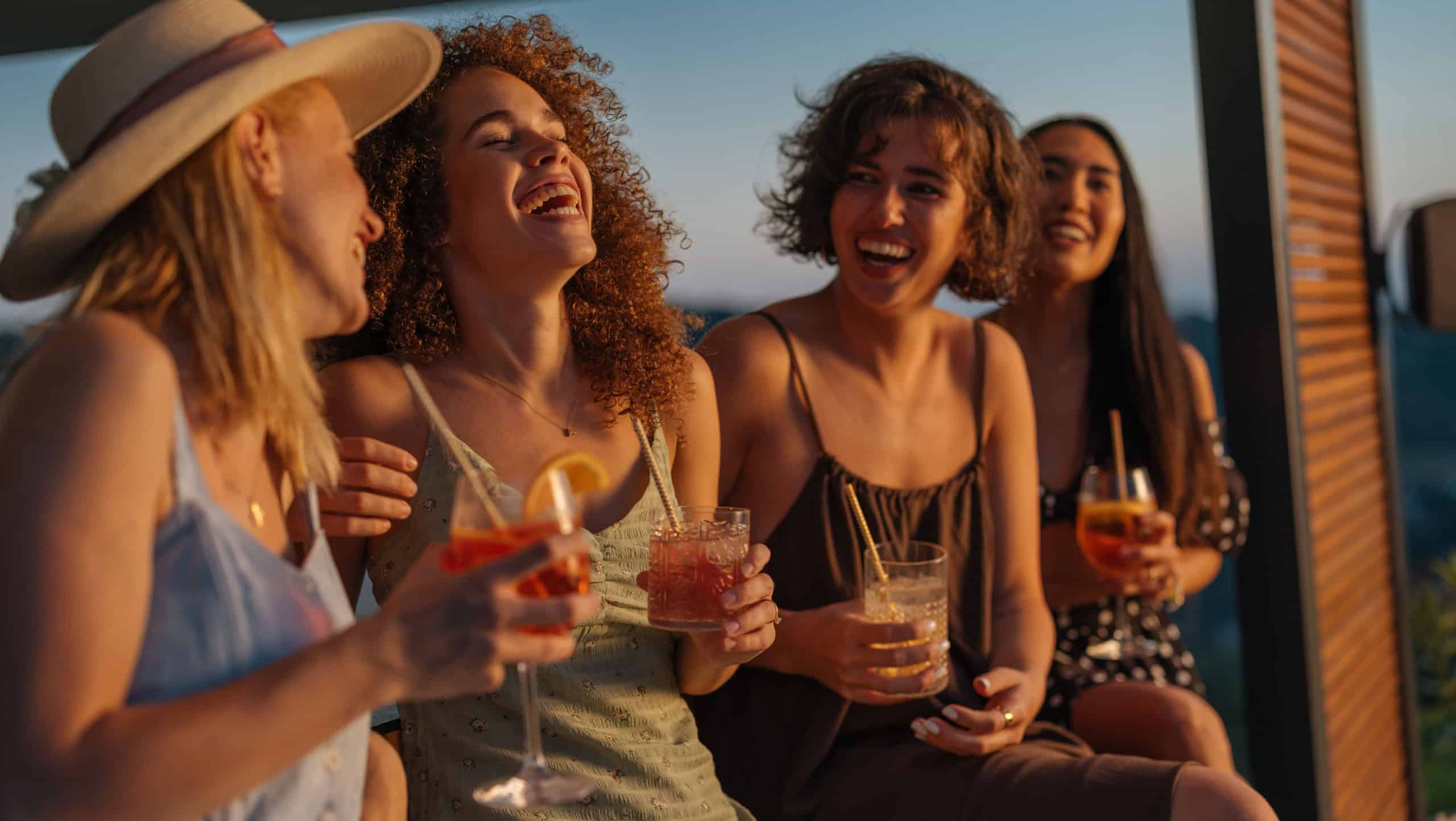 Four female friends enjoying drinks at a rooftop bar, laughing and having fun during a summer evening