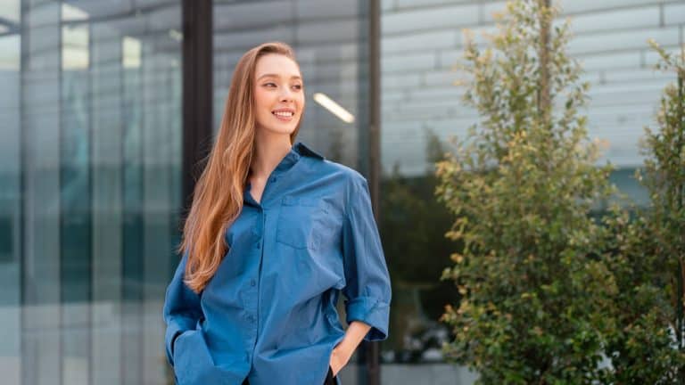 Smiling woman in blue shirt standing with hands in pockets outside modern office, looking away with confidence and calm, reflecting professionalism and smart casual style