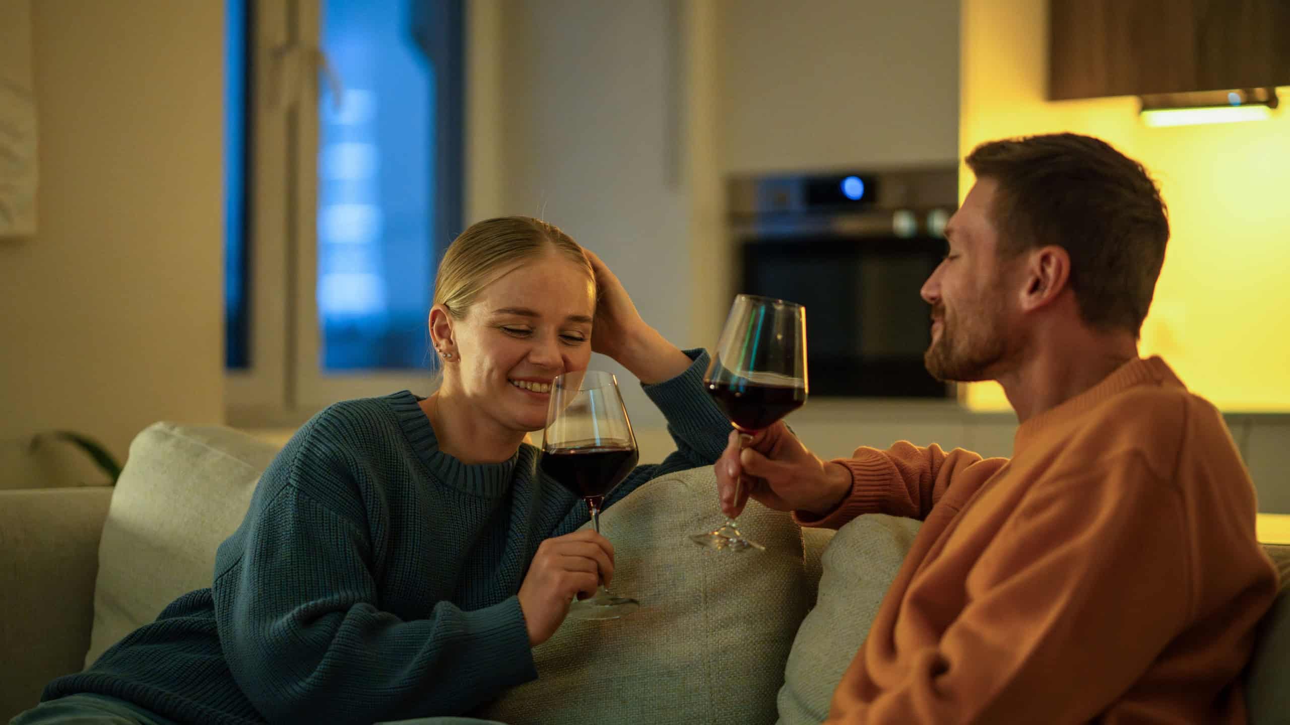 Happy couple enjoying wine together in cozy living room sharing intimate talk on couch at night. Romantic home anniversary date, evening wine tasting together, couple bonding celebration at home