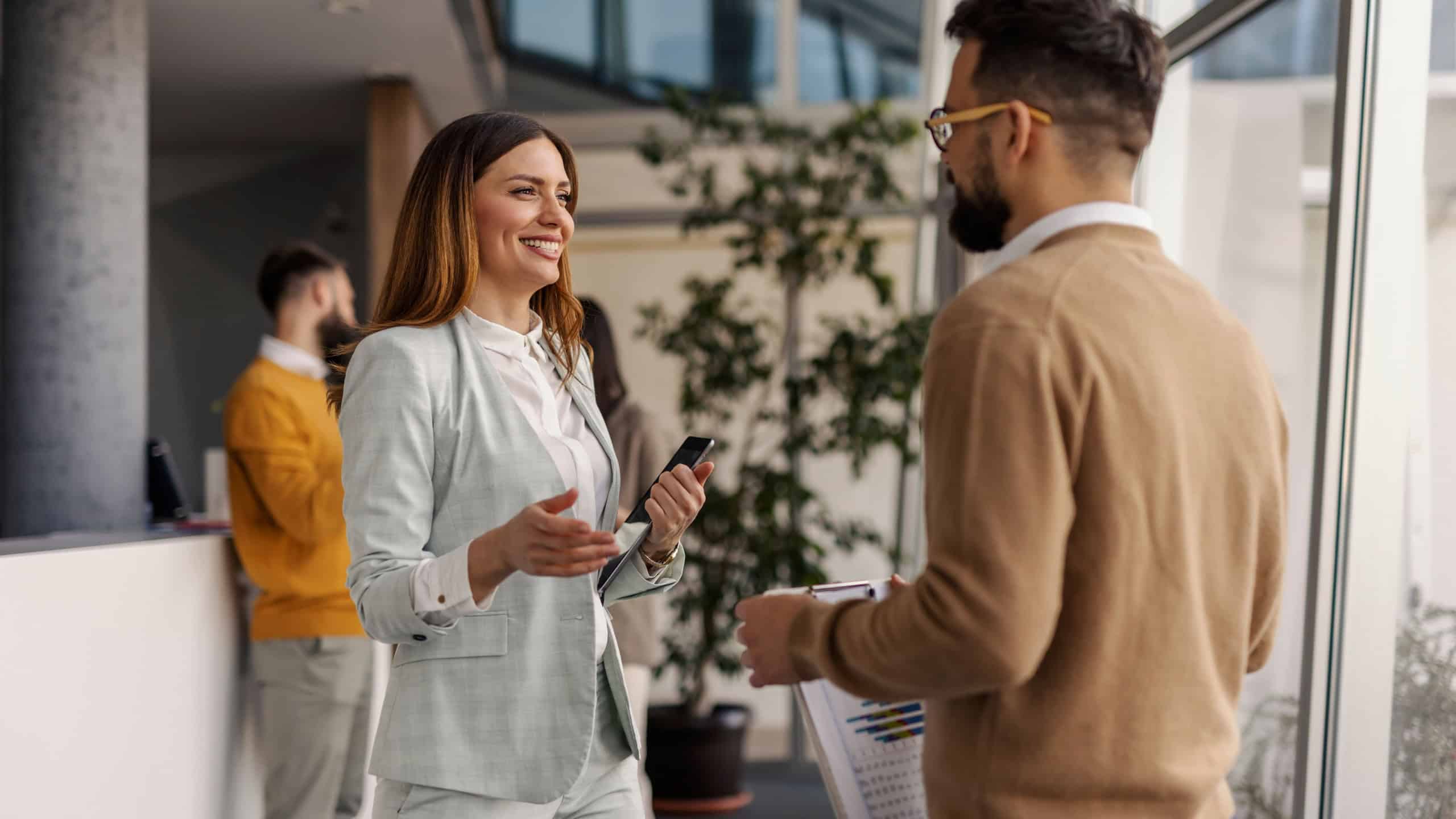 A professional businesswoman and a businessman engage in a friendly discussion in a modern, well-lit office space with large windows and greenery.