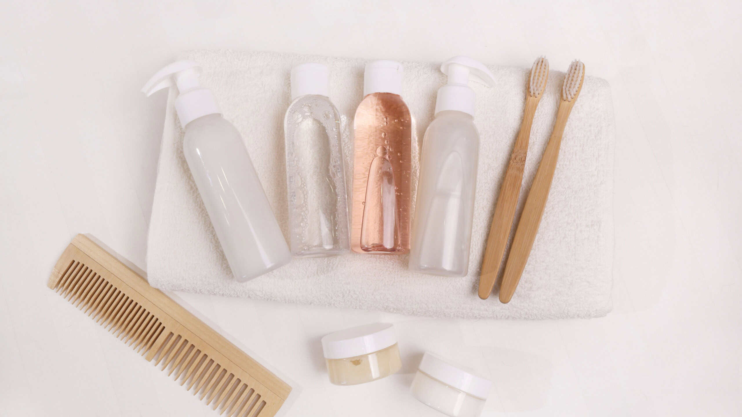 A travel set of toiletries is neatly laid out on a white towel in a hotel.It includes essential personal hygiene items in bottles, wooden toothbrushes and a comb. Top view.