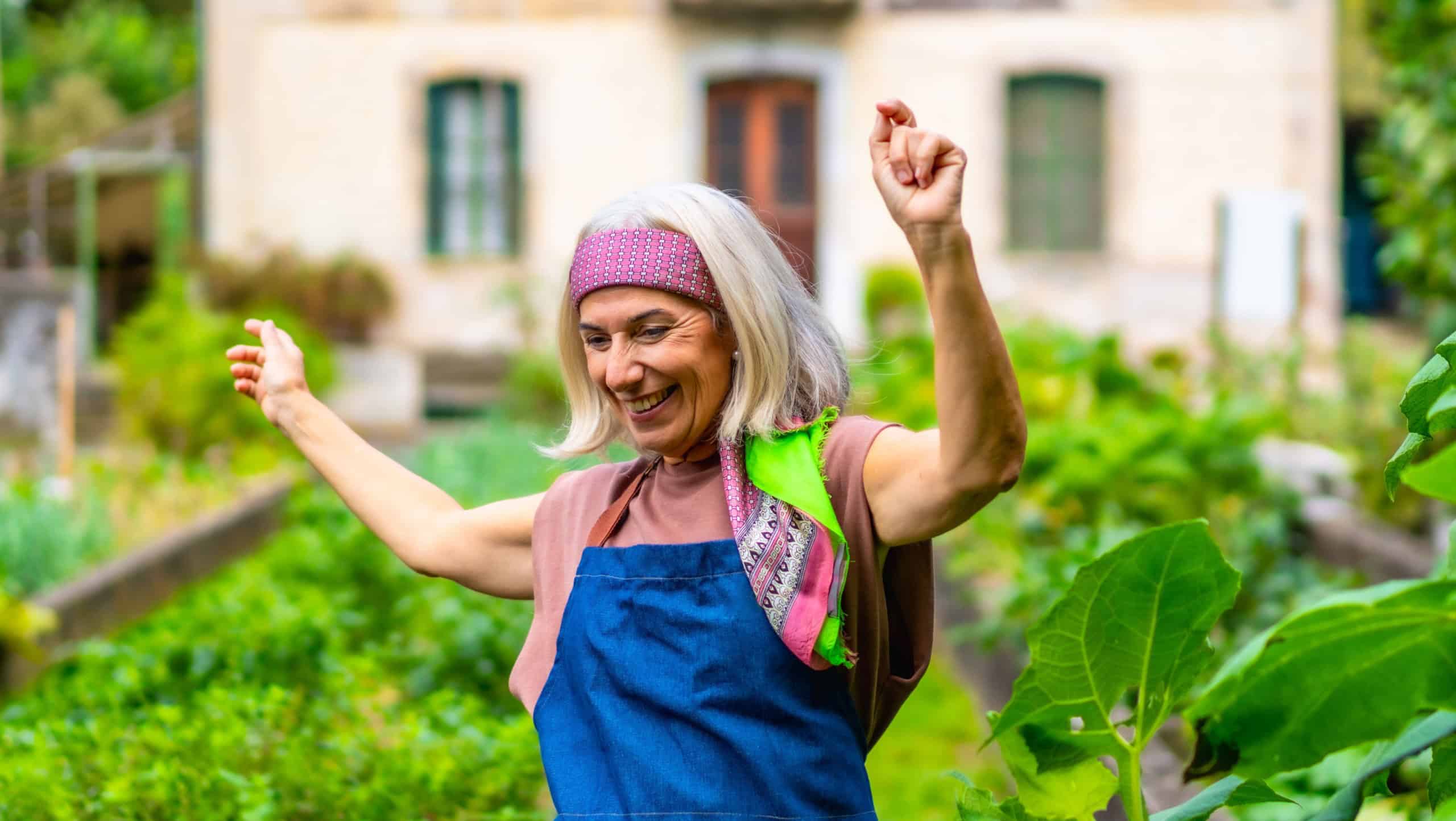 Happy senior woman dancing with arms raised in her home garden, celebrating an active and joyful retirement while cultivating fresh vegetables outdoors