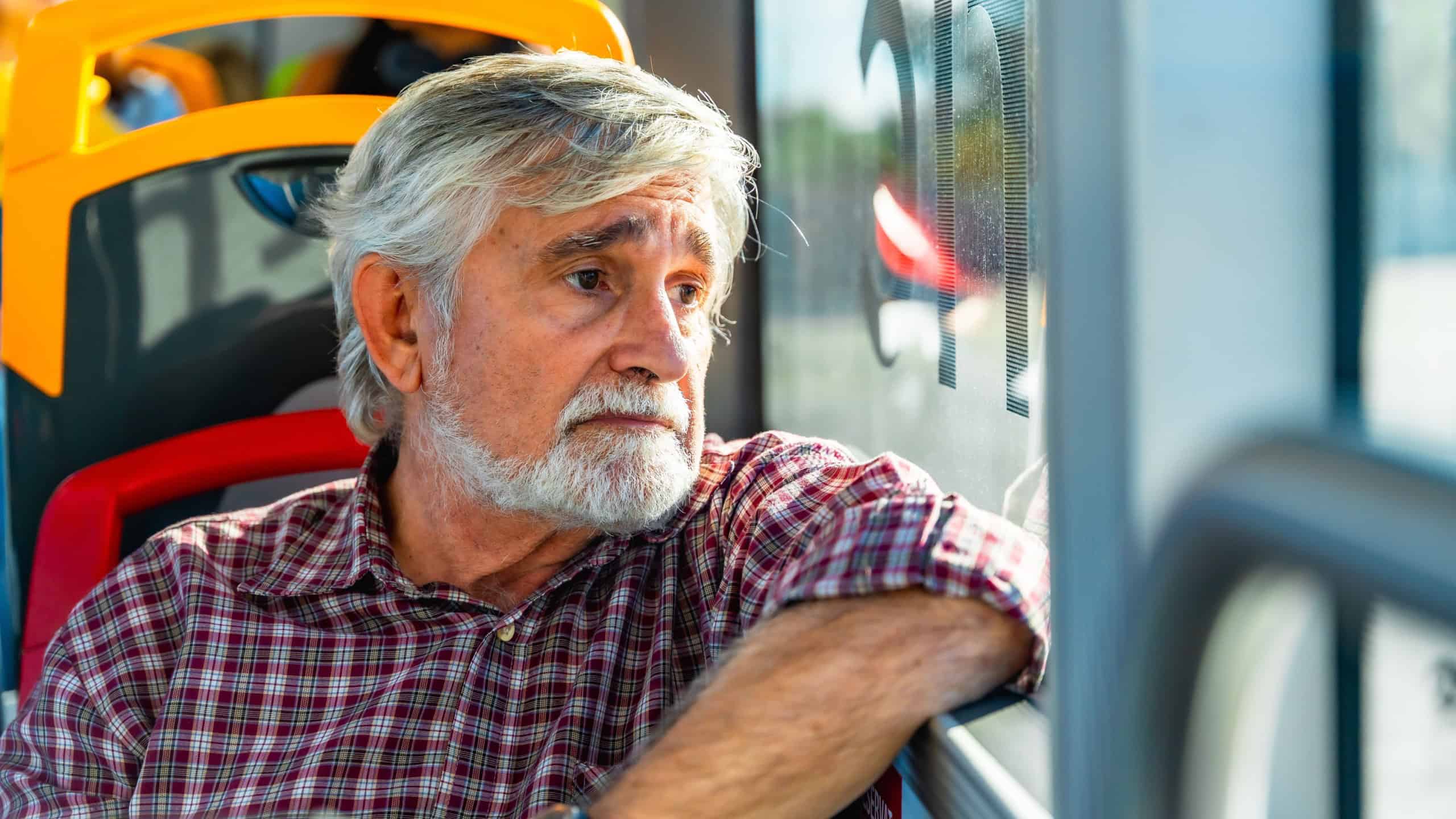 Senior man with gray hair and beard traveling on a public bus, wearing a plaid shirt and looking pensively out of the window, reflecting on his journey