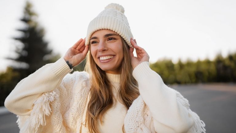 attractive smiling young blond woman walking in winter park having fun wearing warm white knitted sweater, cape and hat, cold season outfit style trend