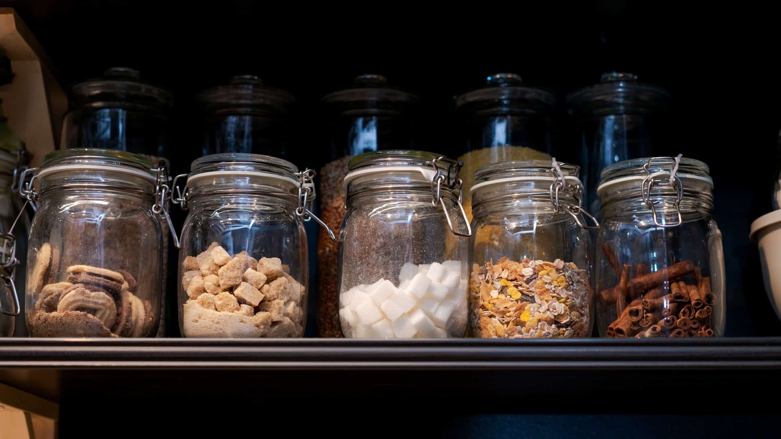 A shelf full of glass jars with various food items inside. The jars are arranged in a row, with some containing sugar, others containing spices, and a few containing cereal