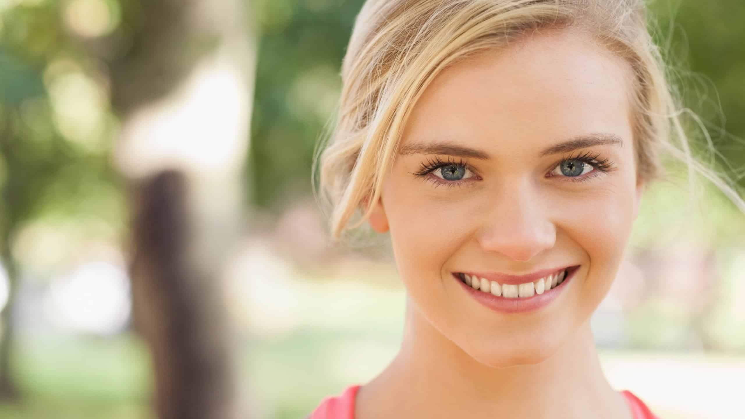 Woman in her 20s smiling posing outdoors in park wearing pink sleeveless top with hair tie. Headshot, outdoor, daylight, bokeh, blonde, female, athletic