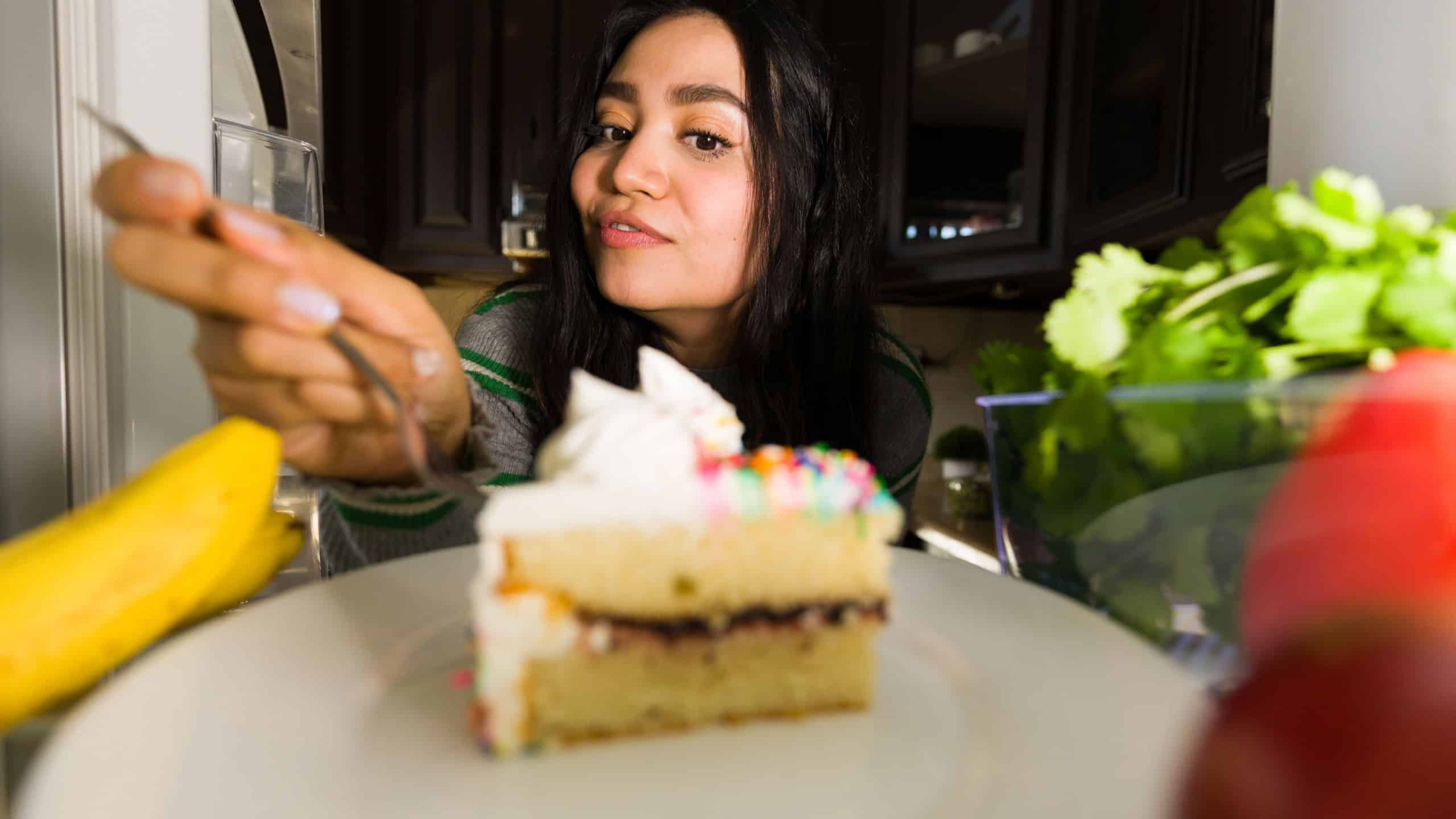 Latin woman reaching inside the fridge, taking a forkful of delicious cake with colorful sprinkles after dark