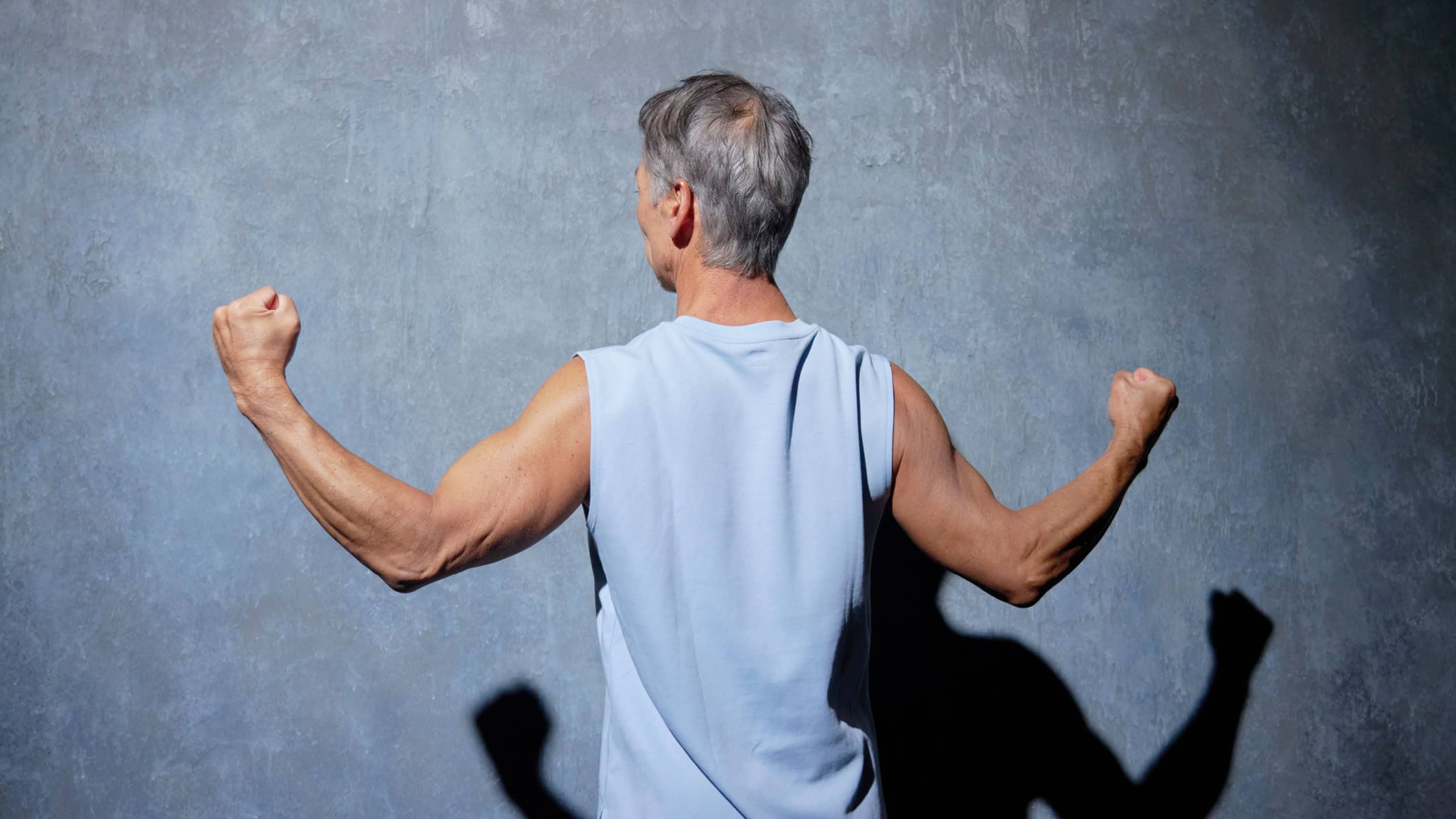 Strong Asian senior man flexing back muscles in sportswear against dark background