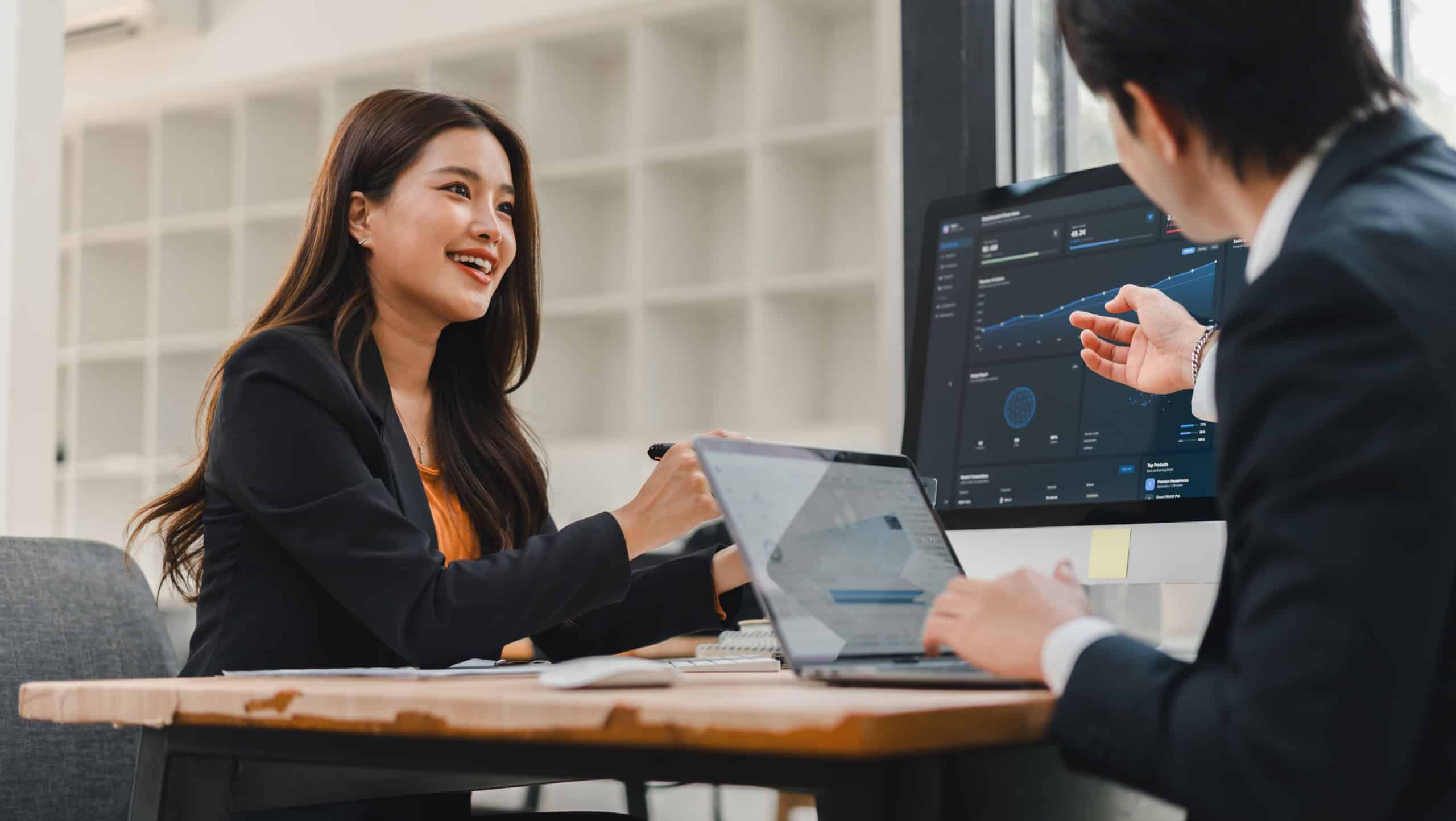 Two Asian professionals discussing business analytics and financial charts on a desktop computer monitor in a modern office.