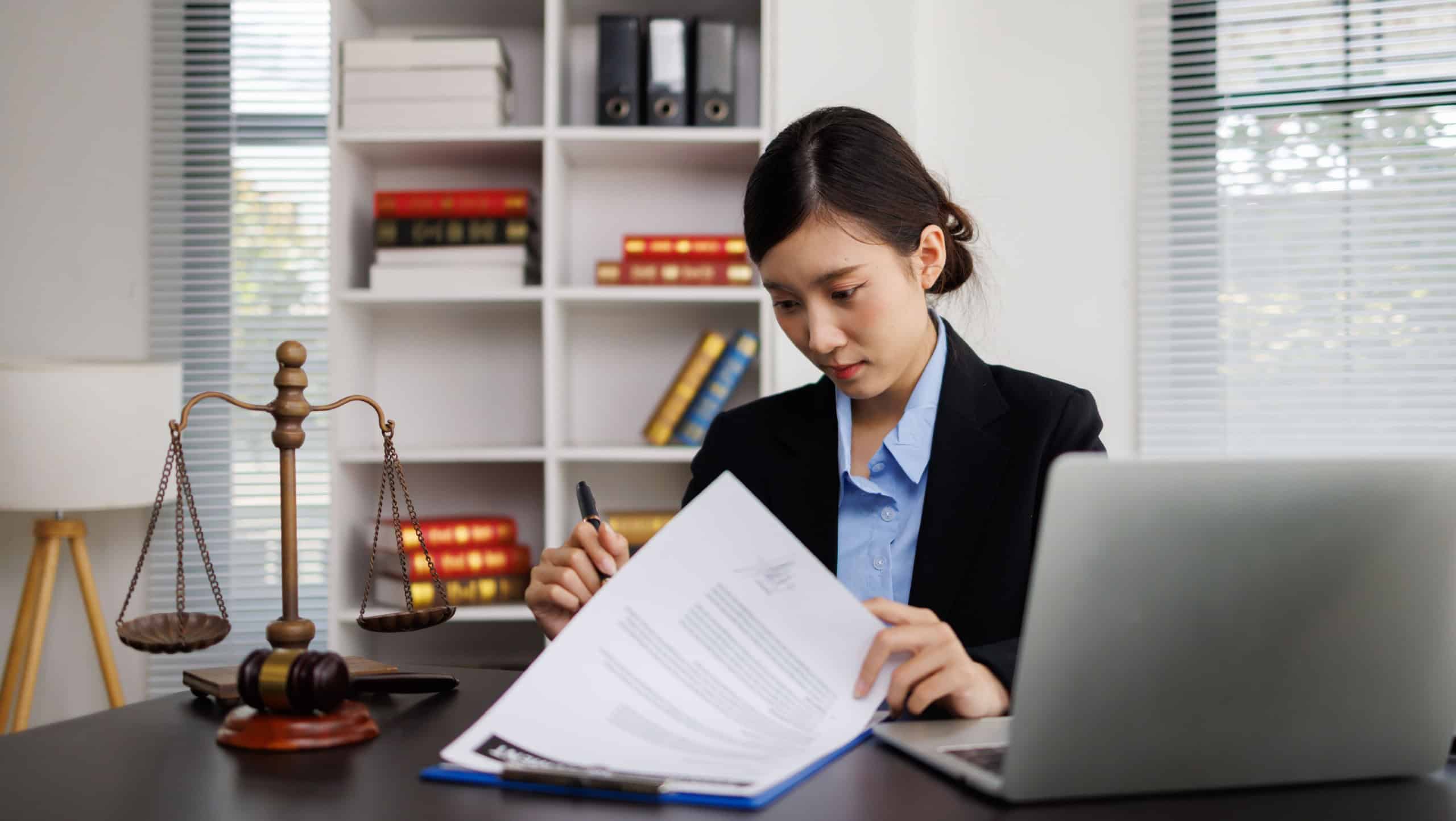 Young Asian female lawyer reviewing legal documents and using a laptop, representing justice, corporate law, and professional consulting in an office setting