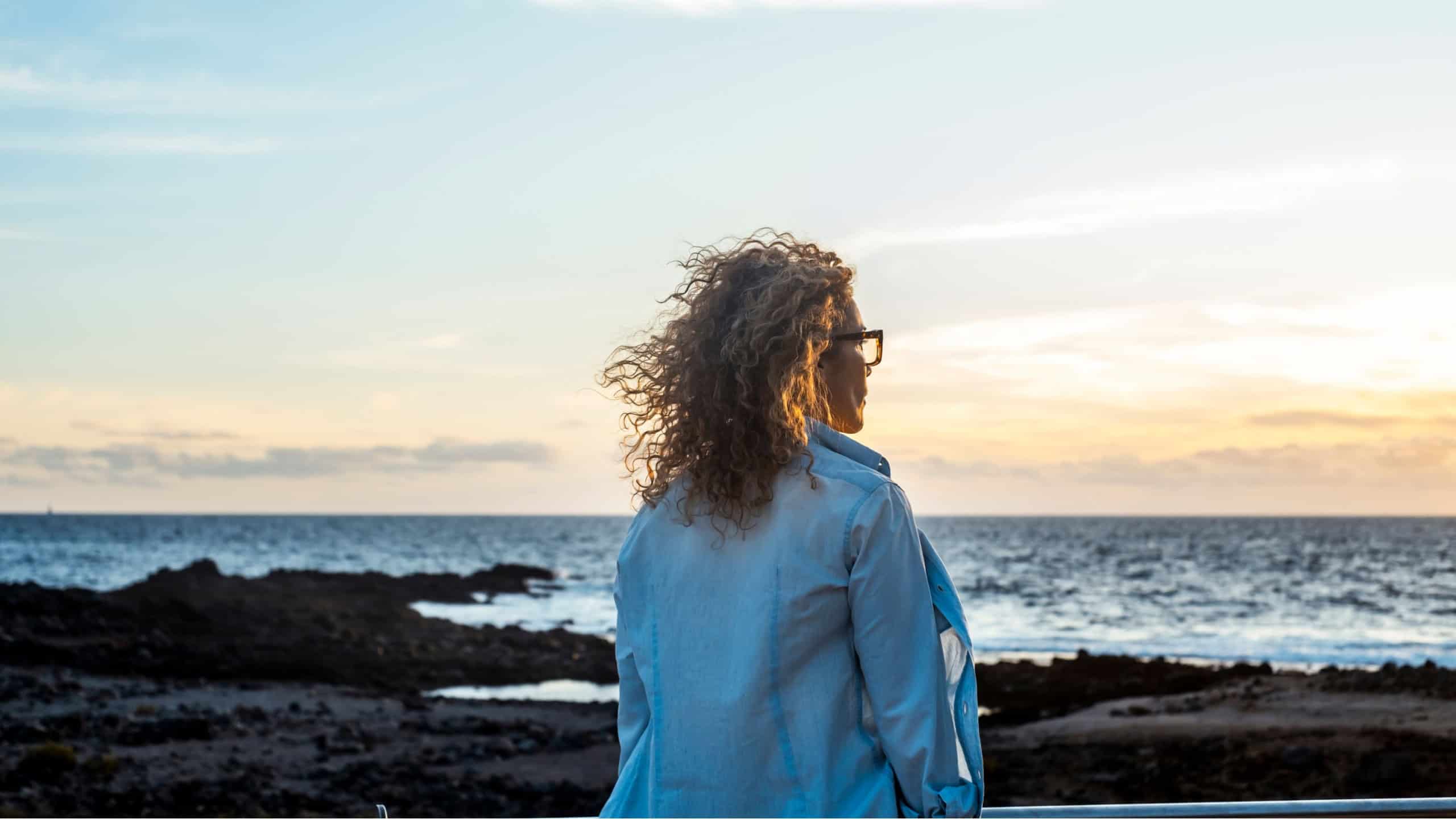 Rear view of mature woman standing on beach looking at sea, feeling melancholic and reflective. Outdoor winter scene with ocean horizon, solitude, memories and emotional lifestyle moment