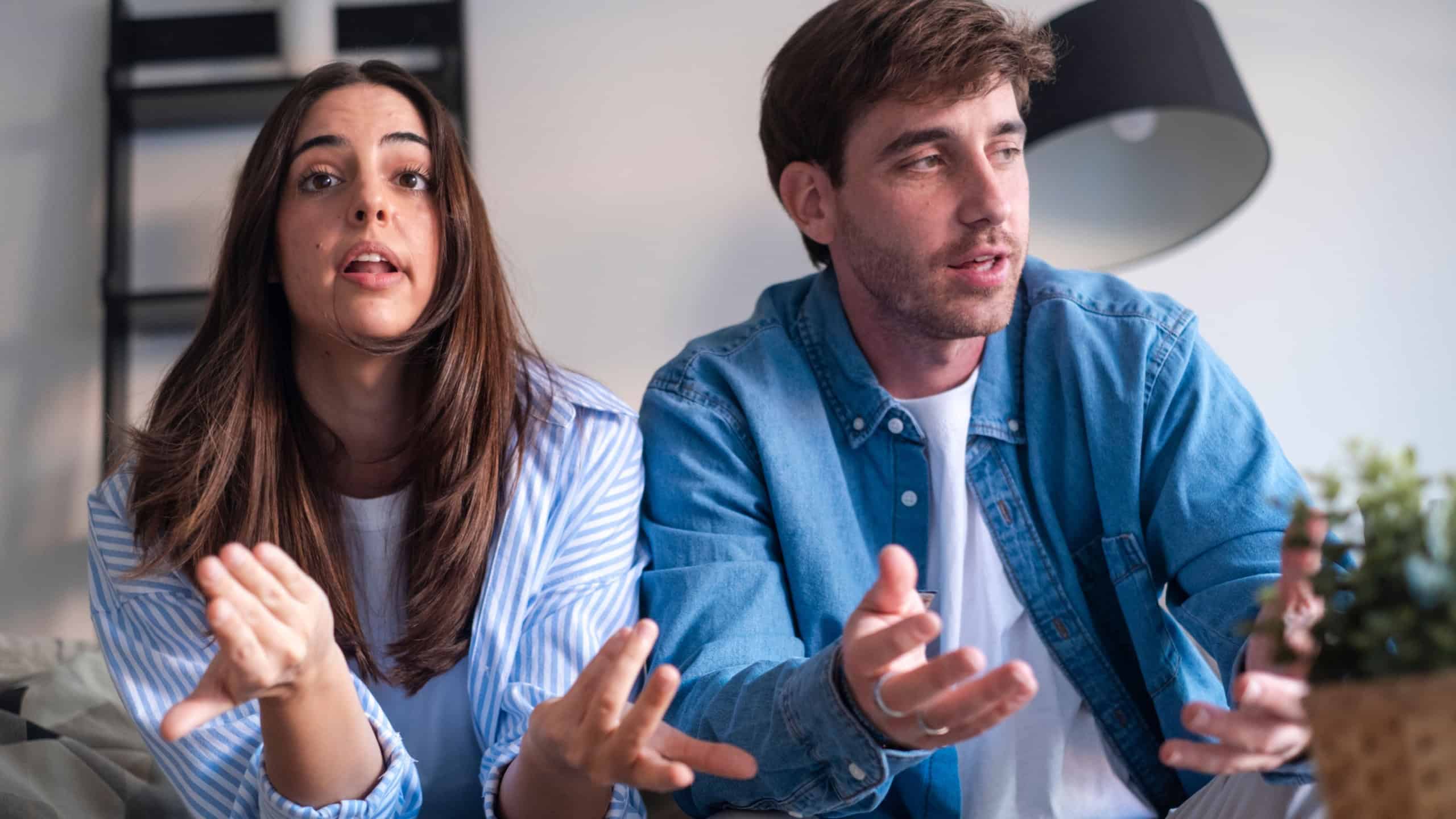 A young couple sits on a couch in their living room having a tense, emotional conversation