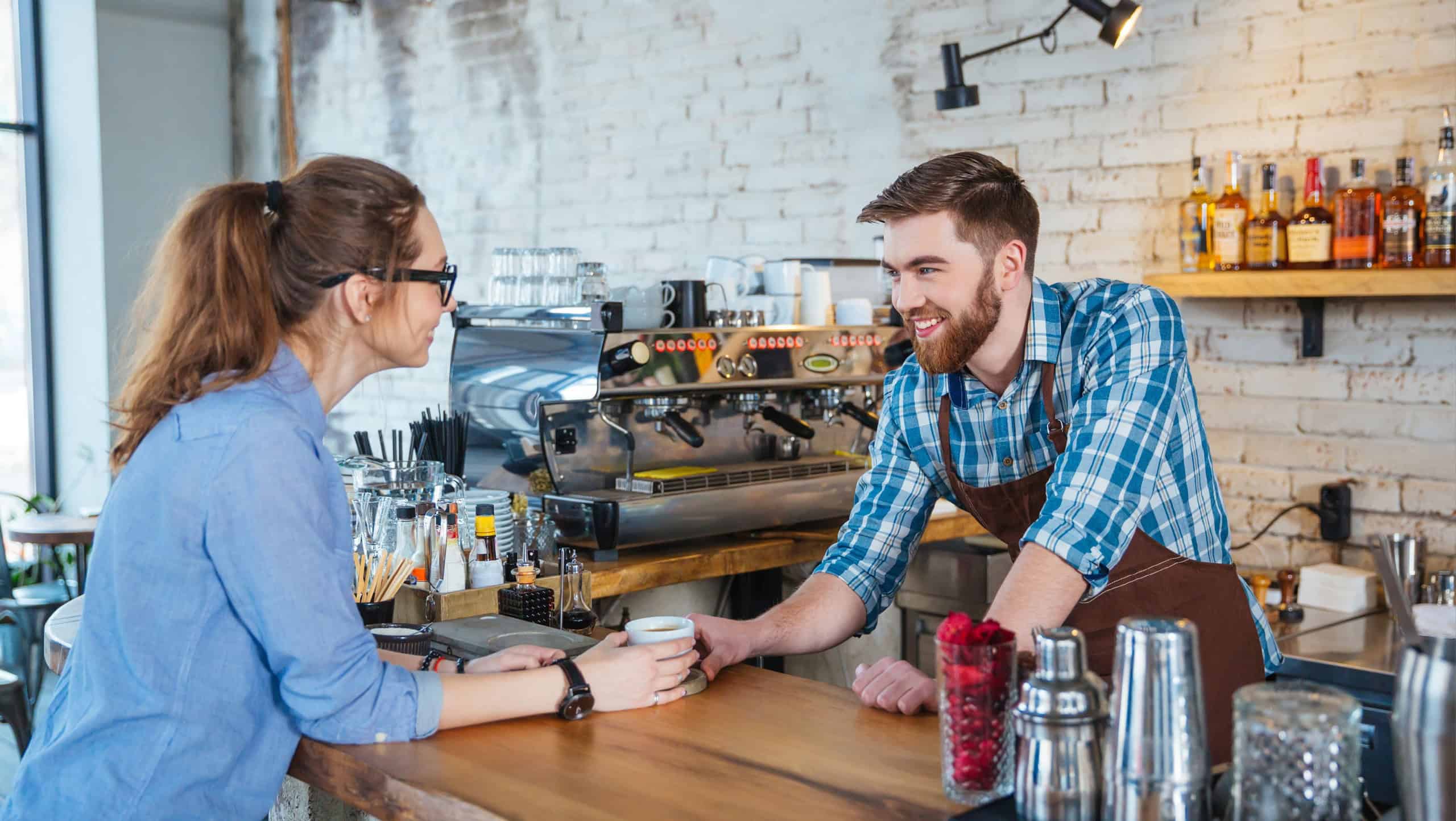 Beautiful young woman in glasses drinking coffee in cafe and talking with handsome barista