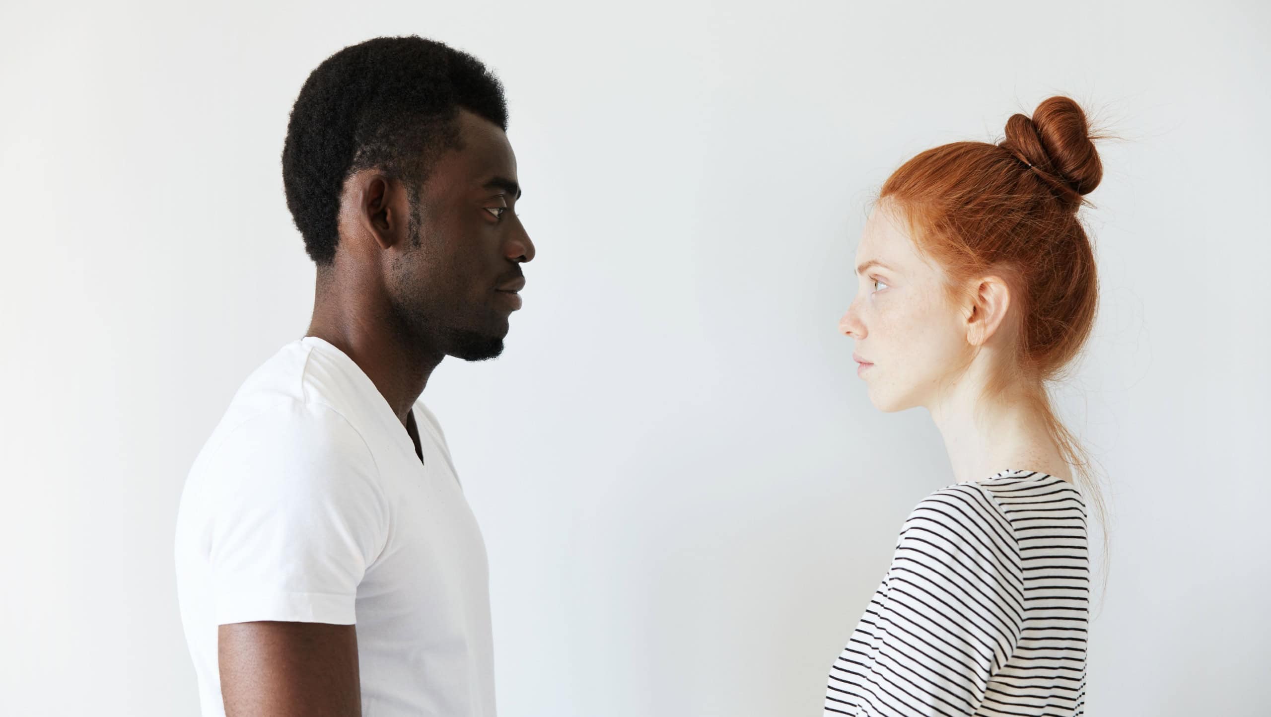Sideways portrait of a couple looking each other in the eyes on white background. Afro man standing still and Caucasian redhead girl as well watching him with serious look like wife on husband.