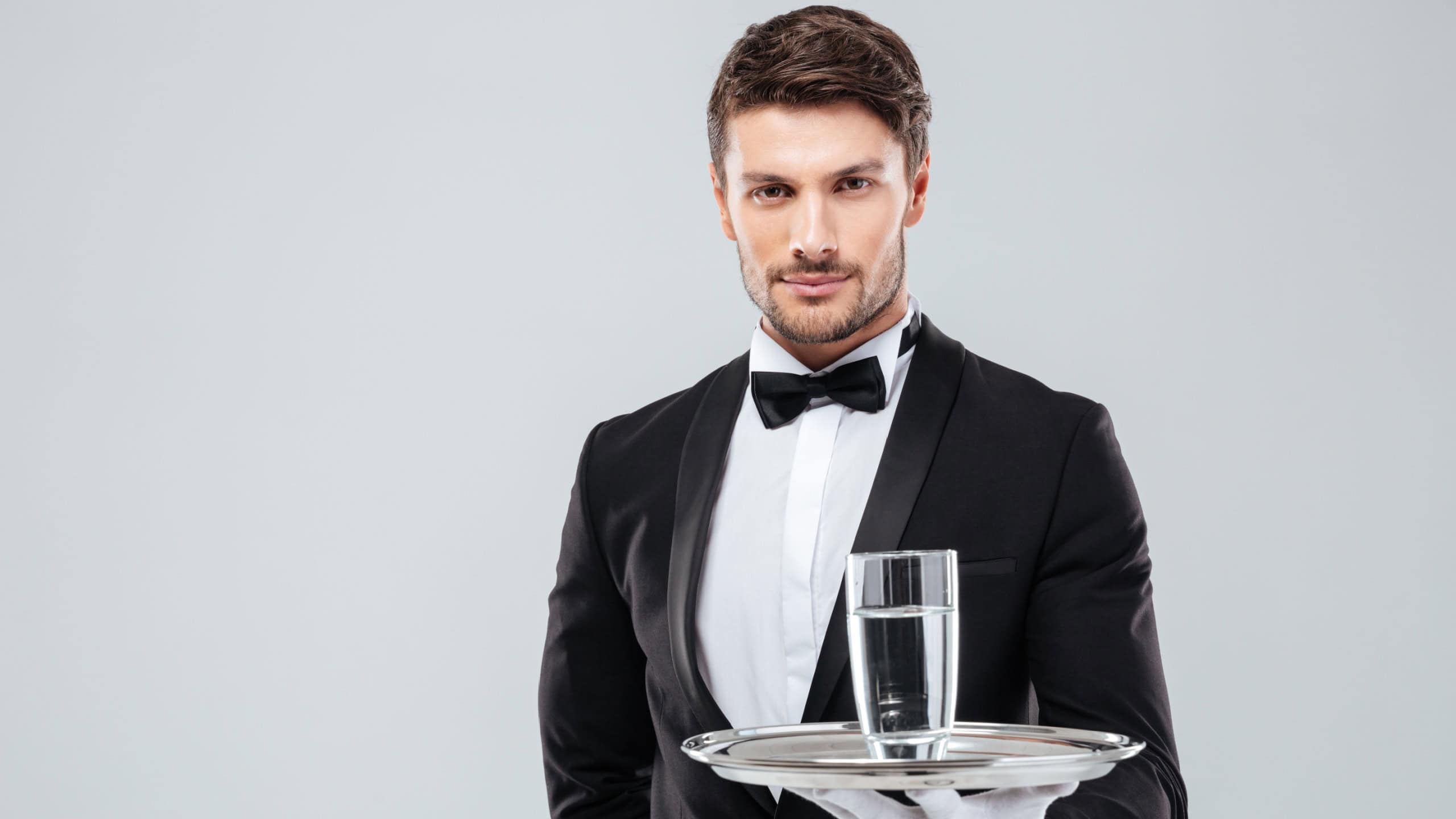 Portrait of waiter in tuxedo and gloves holding glass of water on metal tray