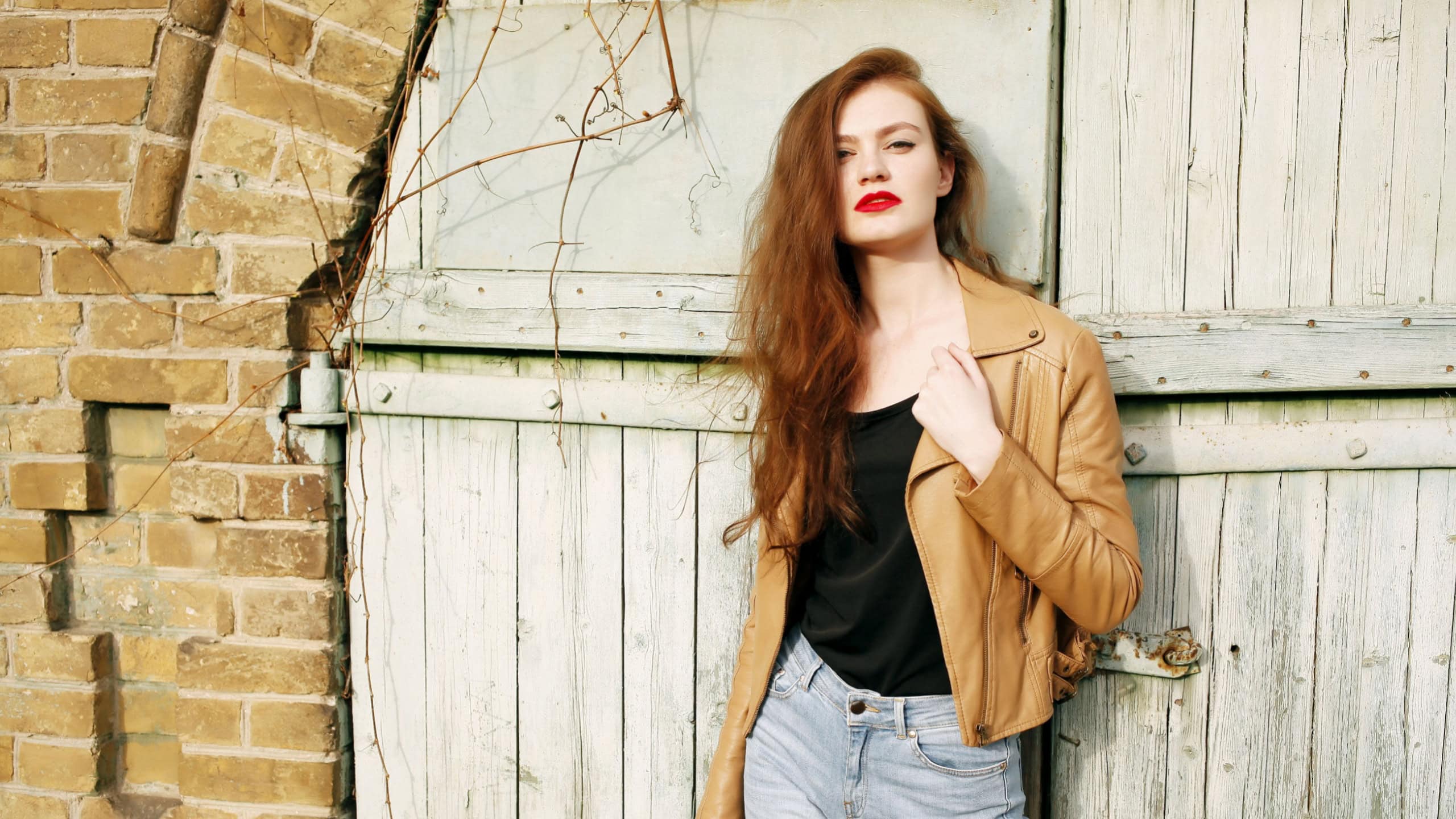 Street fashion concept - closeup portrait of a pretty hipster girl. Wearing brown leather jacket and high waisted jeans. Beautiful autumn woman with red libs and curly hair. Artsy bohemian style.