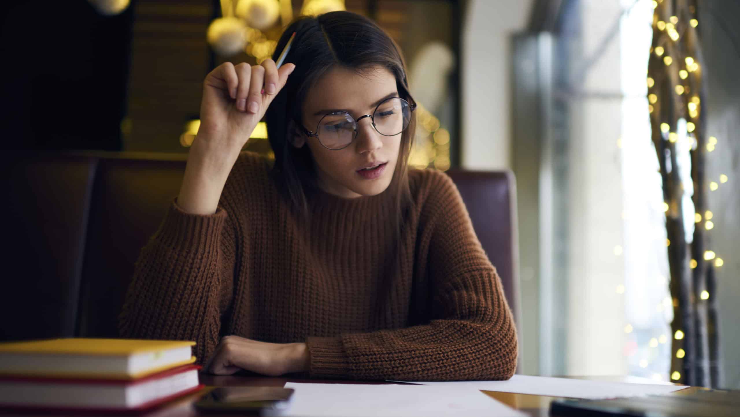 Pensive young female skilled student in glasses and casual outfit checking report correcting mistakes before sending to coach proofreading in coffee shop during break between lessons in university
