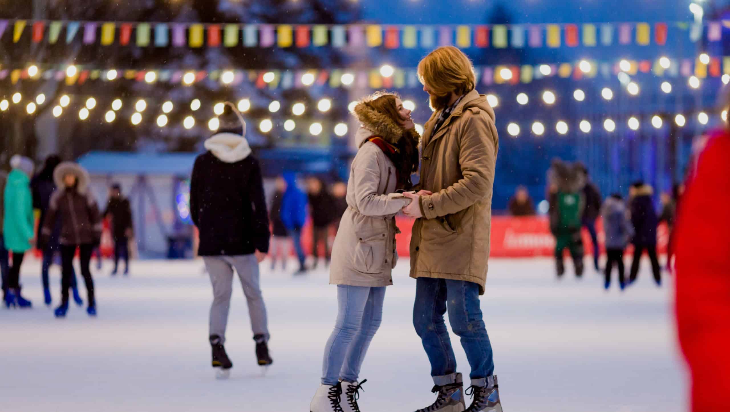 Ice skating rink and lovers together. A pair of young, stylish people in an embrace in a crowd on a city skating rink lit by light bulbs and bright lights. Winter date for Christmas on the ice arena