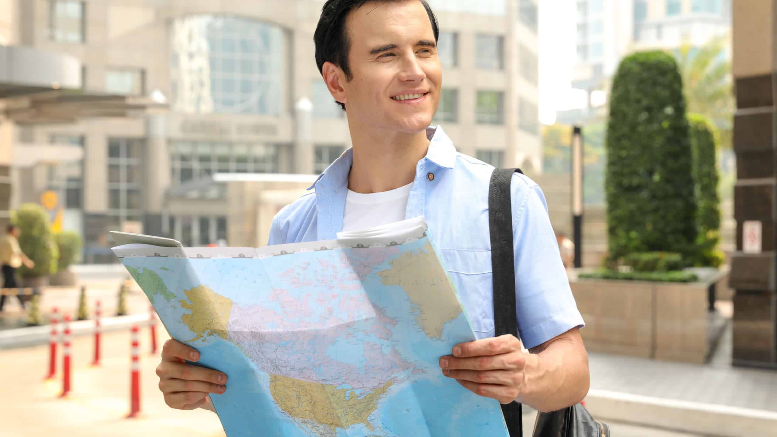 Handsome young traveller in white T-shirt, light blue shirt with world map in hand on city background.Handsome caucasian man,Happy tourists sightseeing city with map,holiday vacation travel concept.