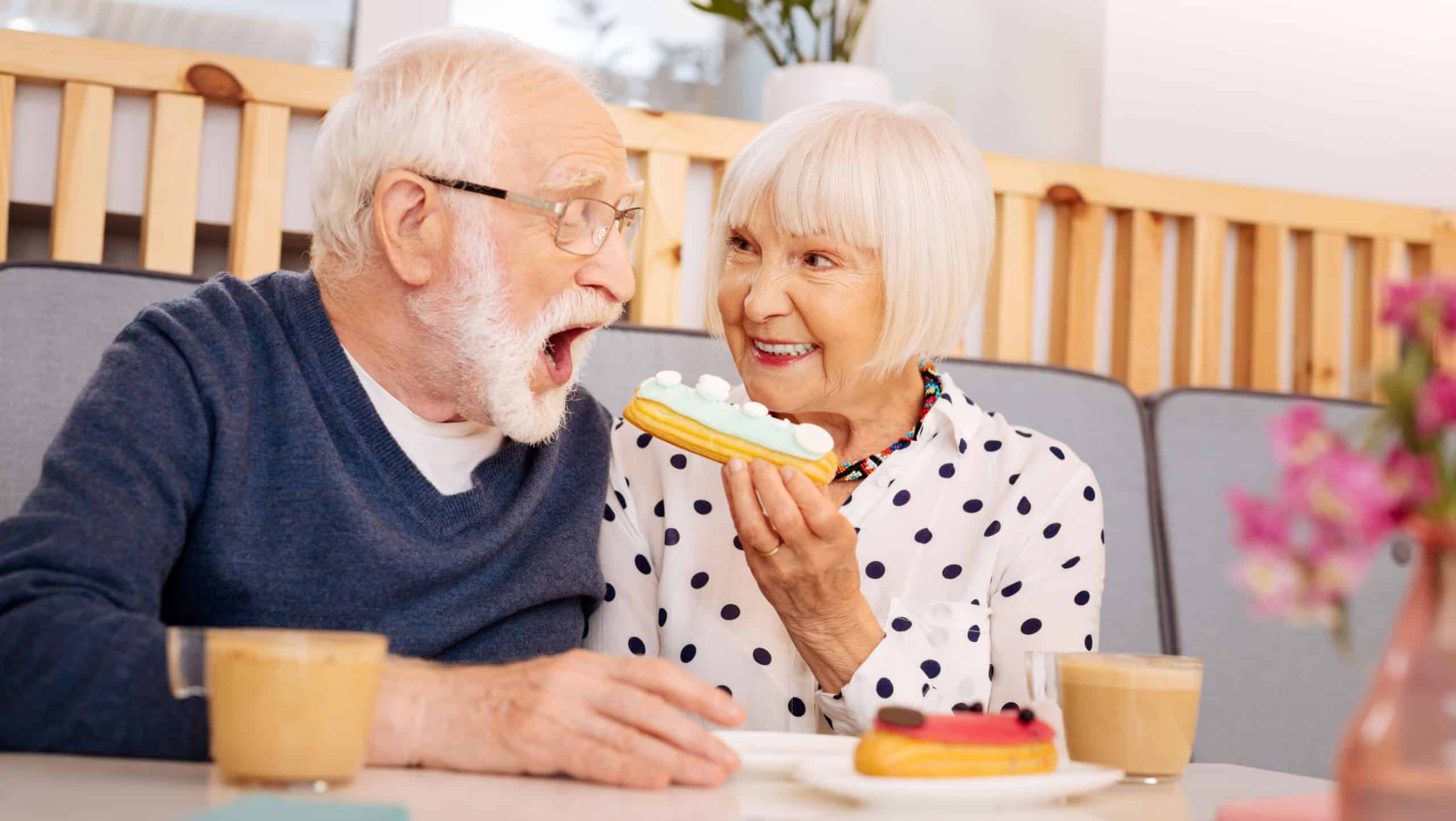 Sweet bonus. Low angle of attractive senior woman holding eclair and senior man opening mouth