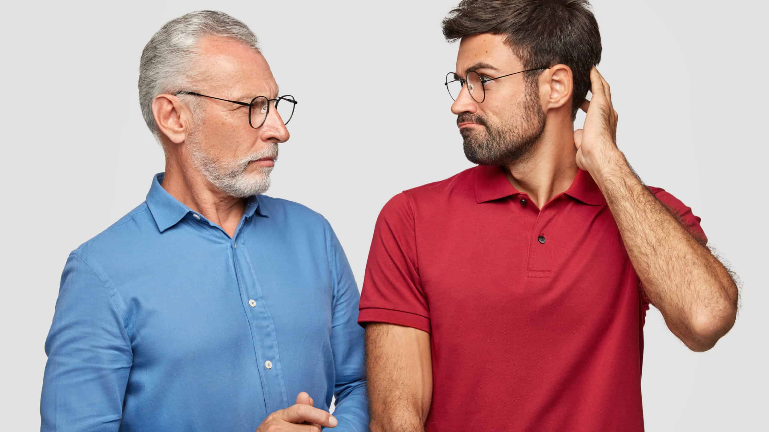 War of generations concept. Displeased bearded mature father and son look angrily at each other, have argue, cant find common solution, pose against white studio background.