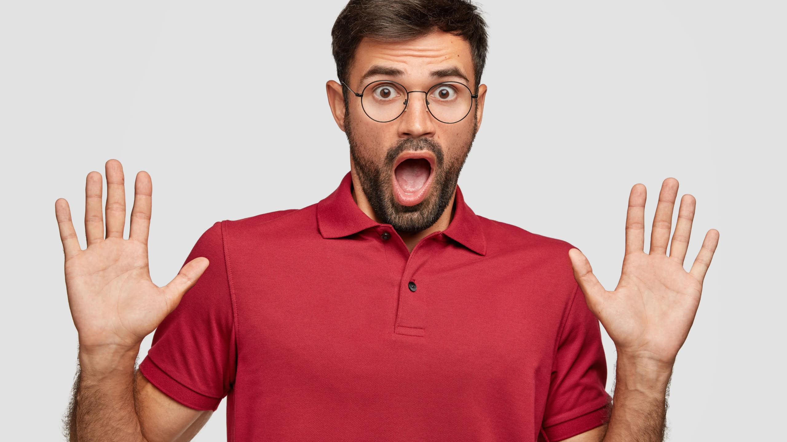 Mad man keeps palms in front, has shocked expression, opens mouth widely, has terrific look, exlaims he is not guilty, dressed in casual red t shirt stands over white background. Oh no, it cant be so!
