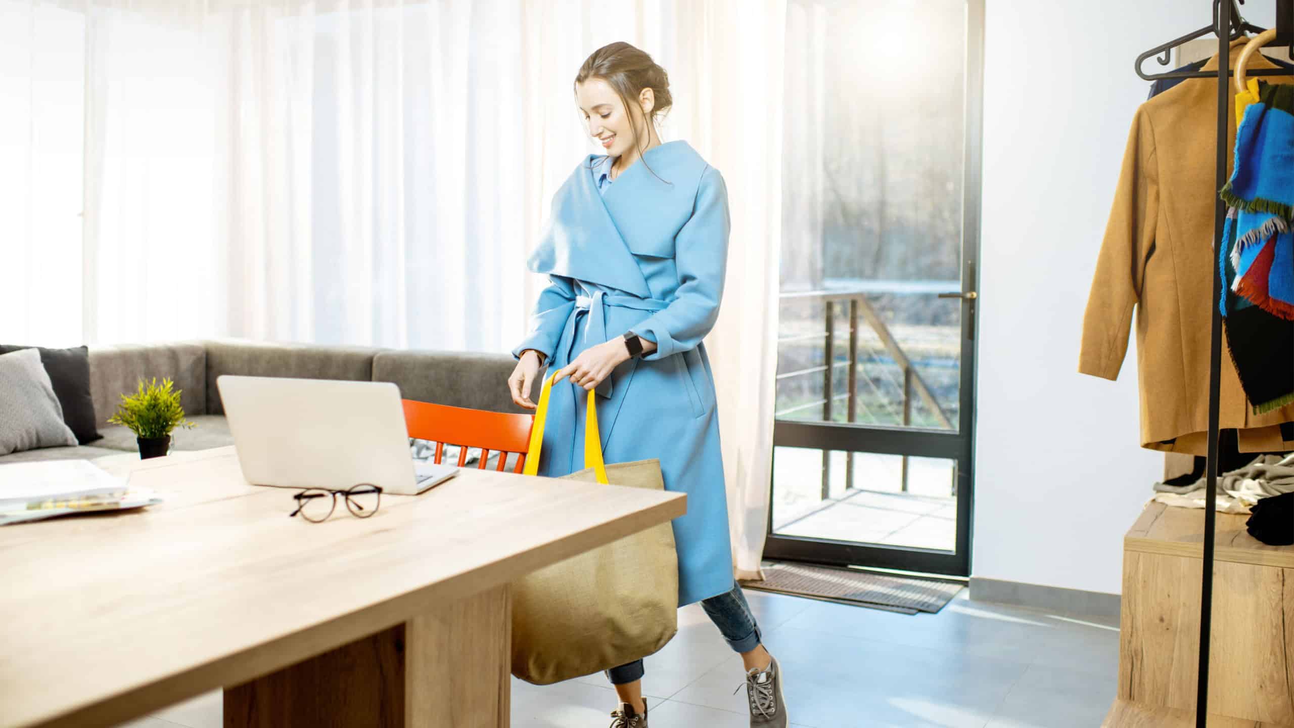 Young stylish woman in blue coat coming home with shopping bag, walking in the living room of the modern apartment