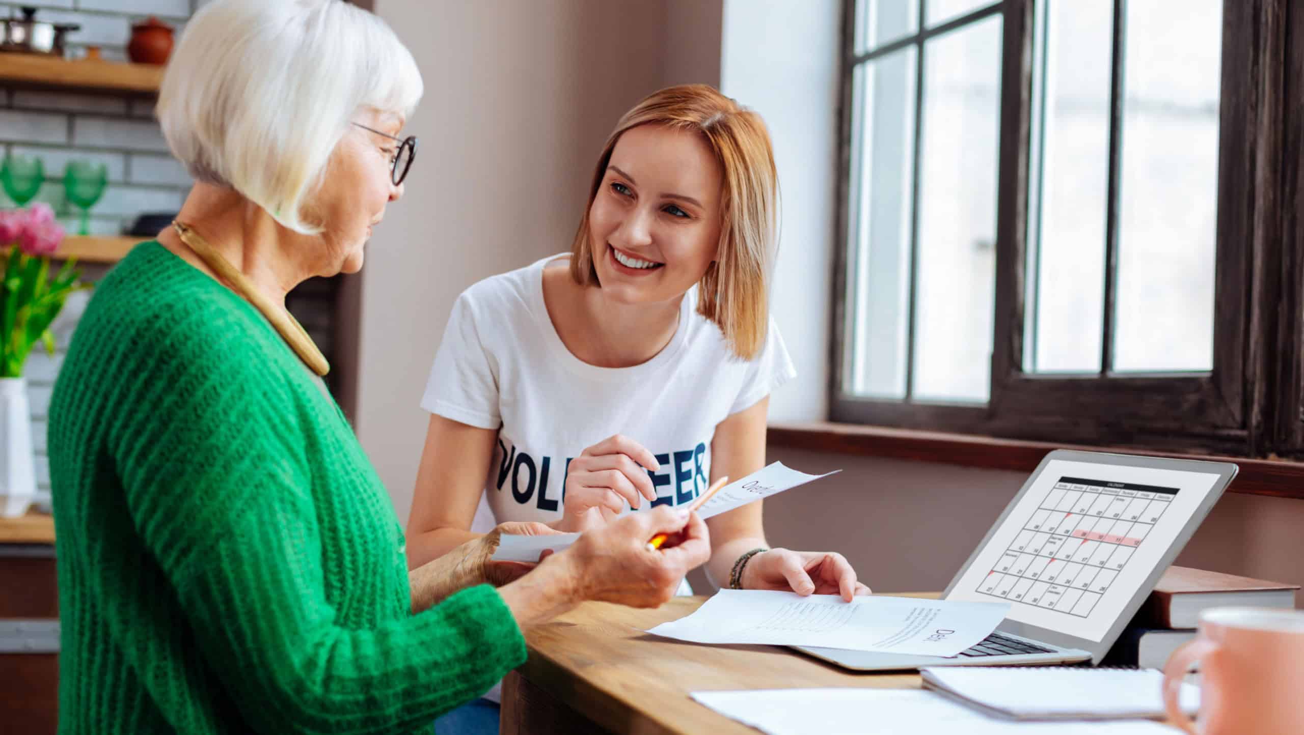 Considering loan payment calendar. Elegant silver-haired retired dame in bright green cardigan looking at loan payment date on laptop calendar with young-adult smiling blond woman helping her