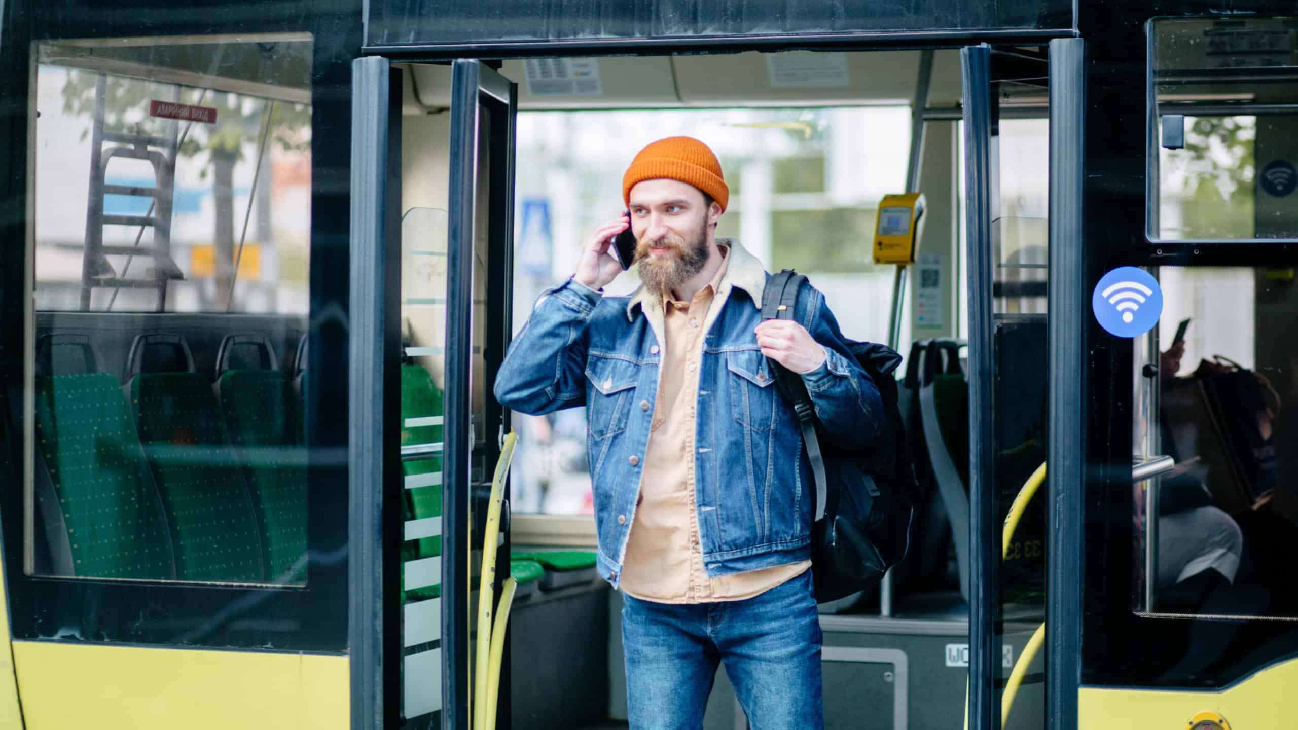 Portrait of happy young hipster american travel man in blue denim jacket and orange hat with bag getting off bus talking on phone on the stop.