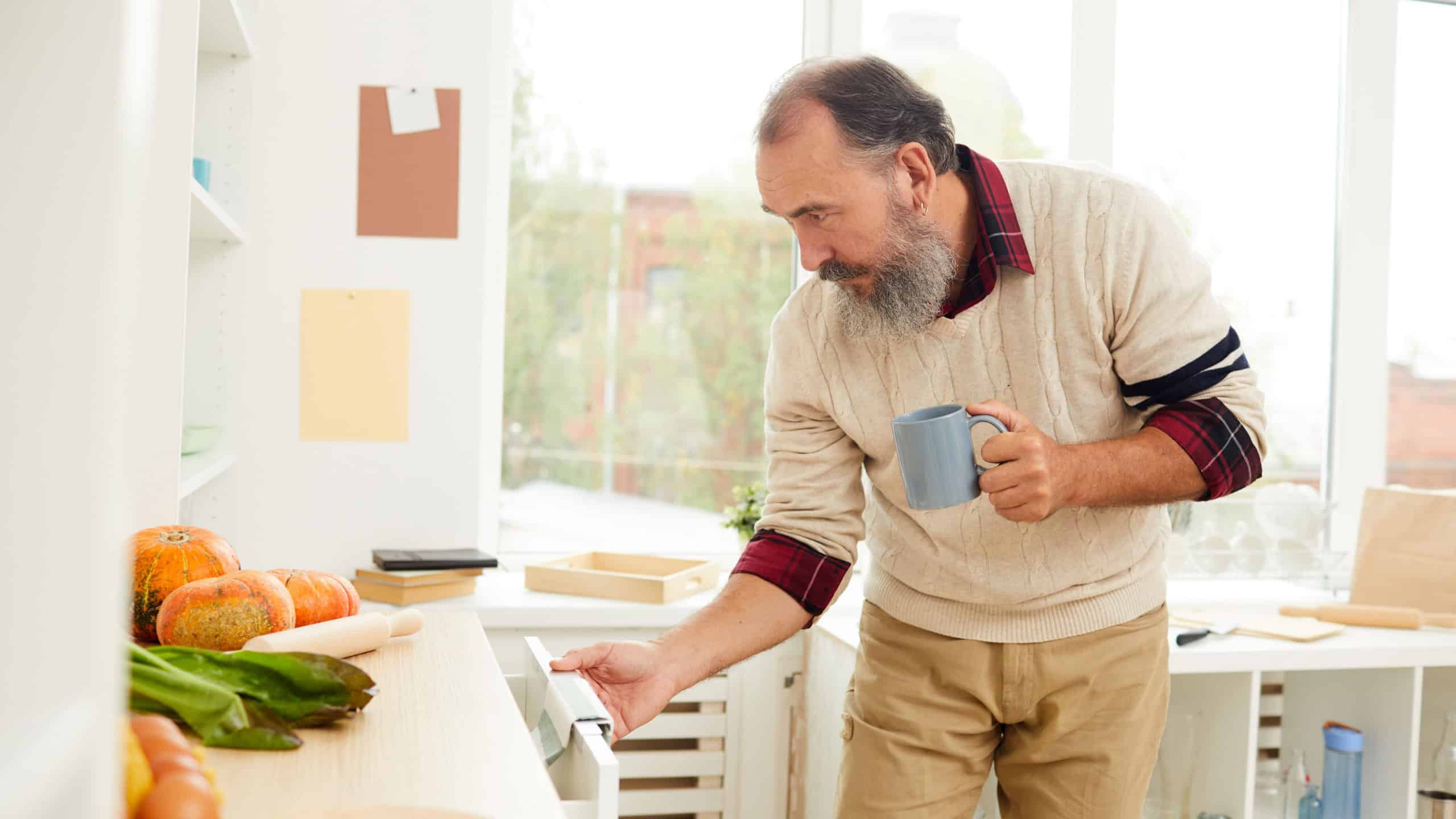 Side view portrait of bearded senior man opening kitchen cupboards while looking for breakfast in morning