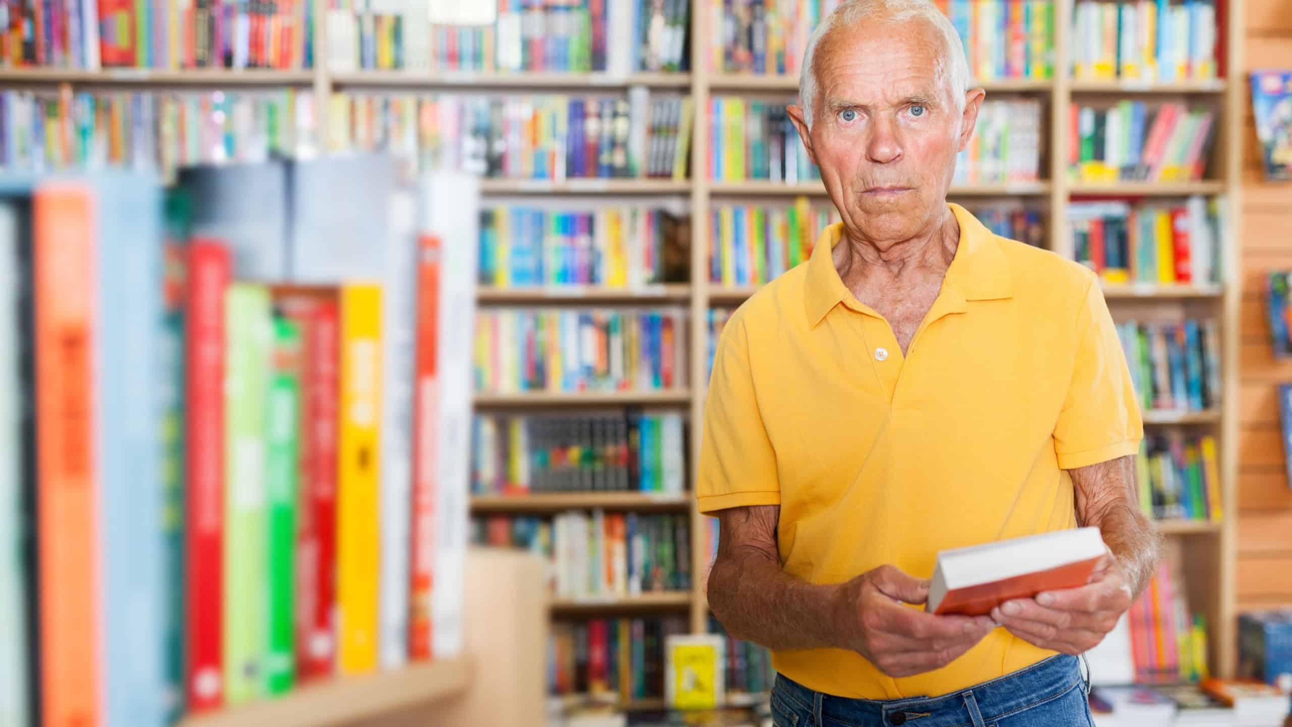 Pensive mature man standing with book in library on background with colorful bookshelves
