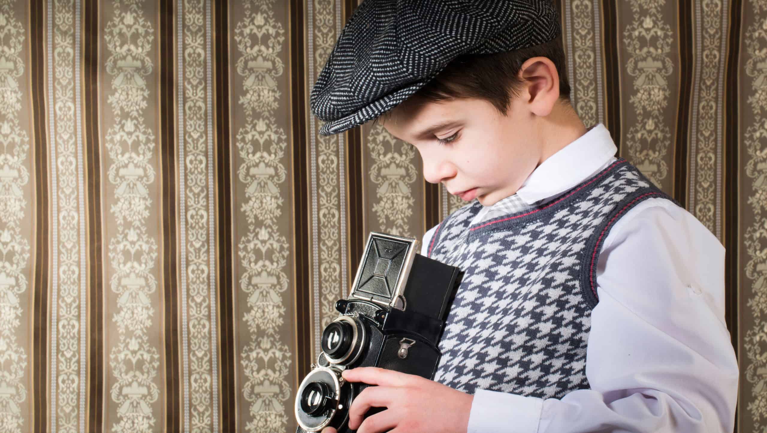 Boy with vintage camera. Vintage clothes. Kid photographer on gothic ornamental wallpaper background. Taking pictures with old camera.