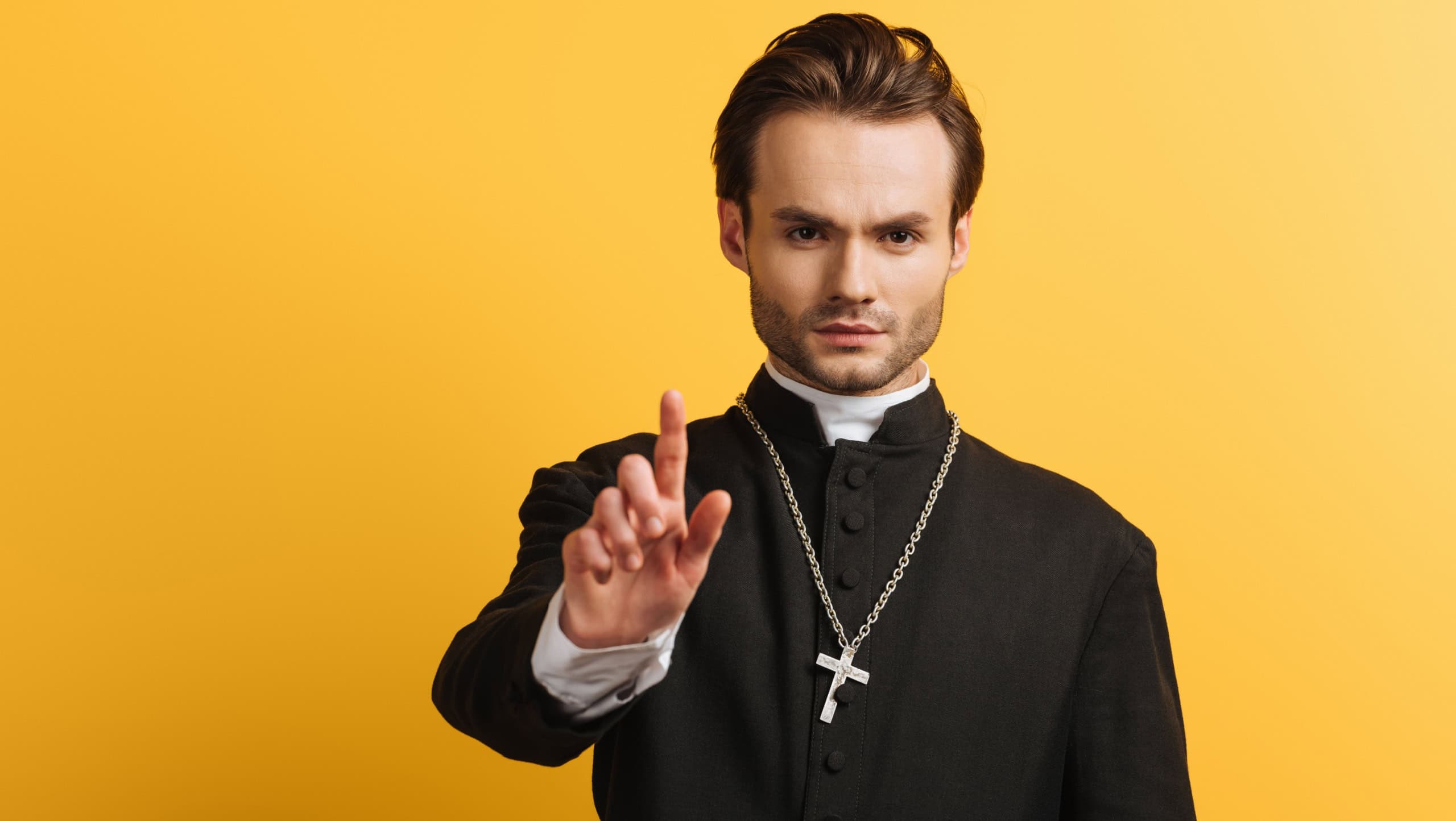 serious catholic priest showing warning gesture isolated on yellow