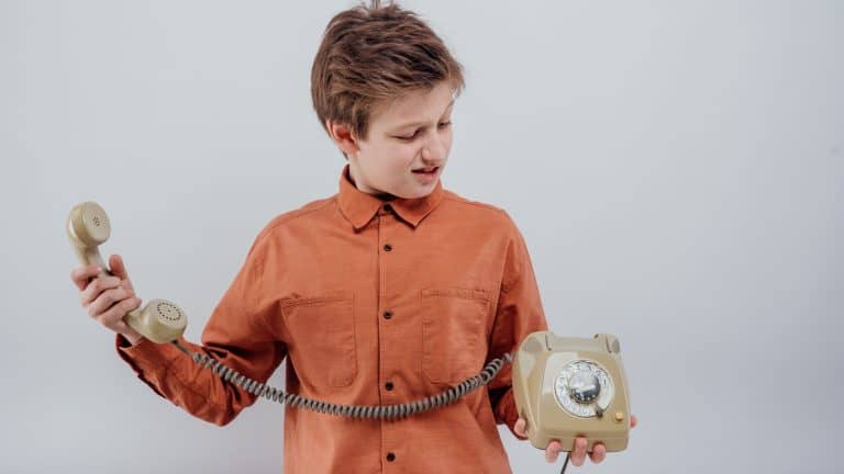 Dissatisfied surprised preteen boy, in orange formal shirt holding and looking at retro disk phone with aversion and misunderstand against gray background in light modern studio