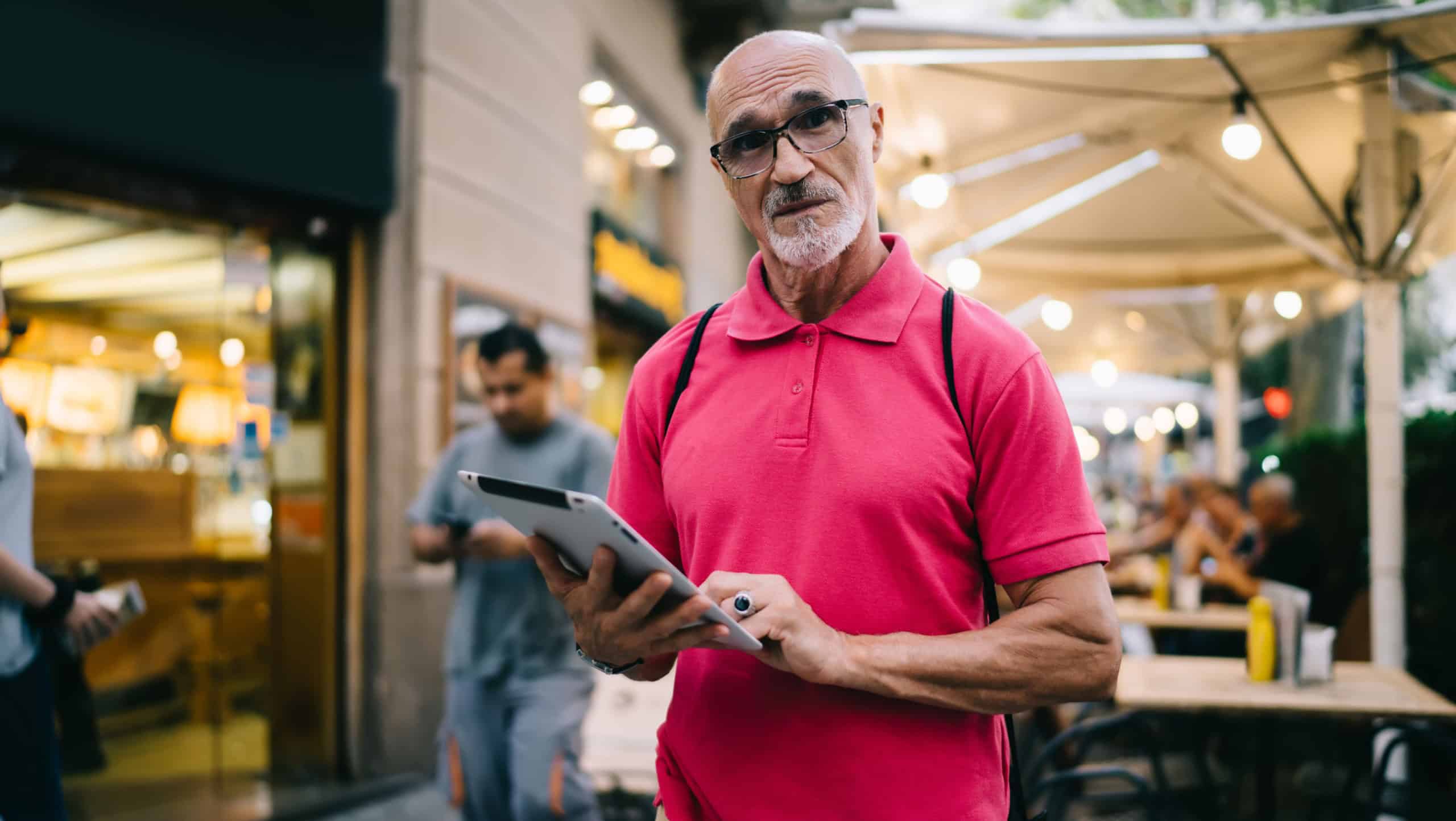 Confused bearded aged man in casual clothes and eyeglasses looking at camera and searching direction on tablet against outdoor cafe