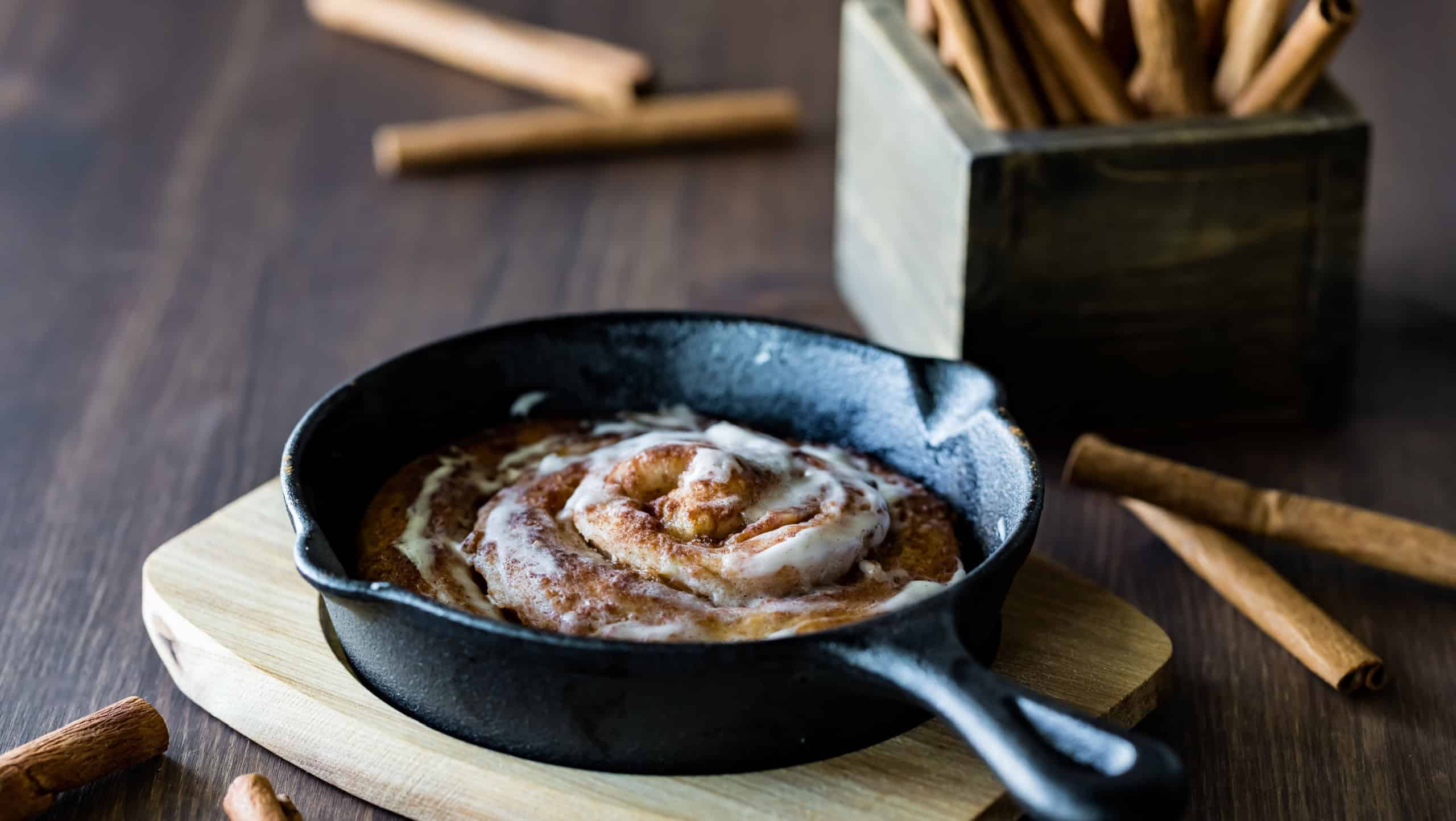 Cinnamon bun baked in a cast iron frypan with a box of cinnamon sticks in behind.