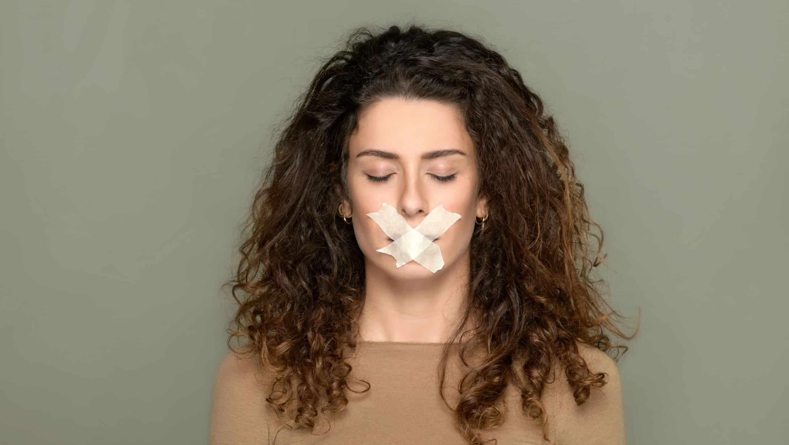 Young woman with closed eyes and her mouth taped shut in a concept of censorship of vision and free speech over a grey studio background