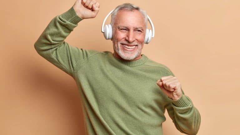 Carefree retired bearded man listens music and dances actively has happy mood smiles toothily wears white wireless headphones on ears dressed in casual sweater isolated over brown background