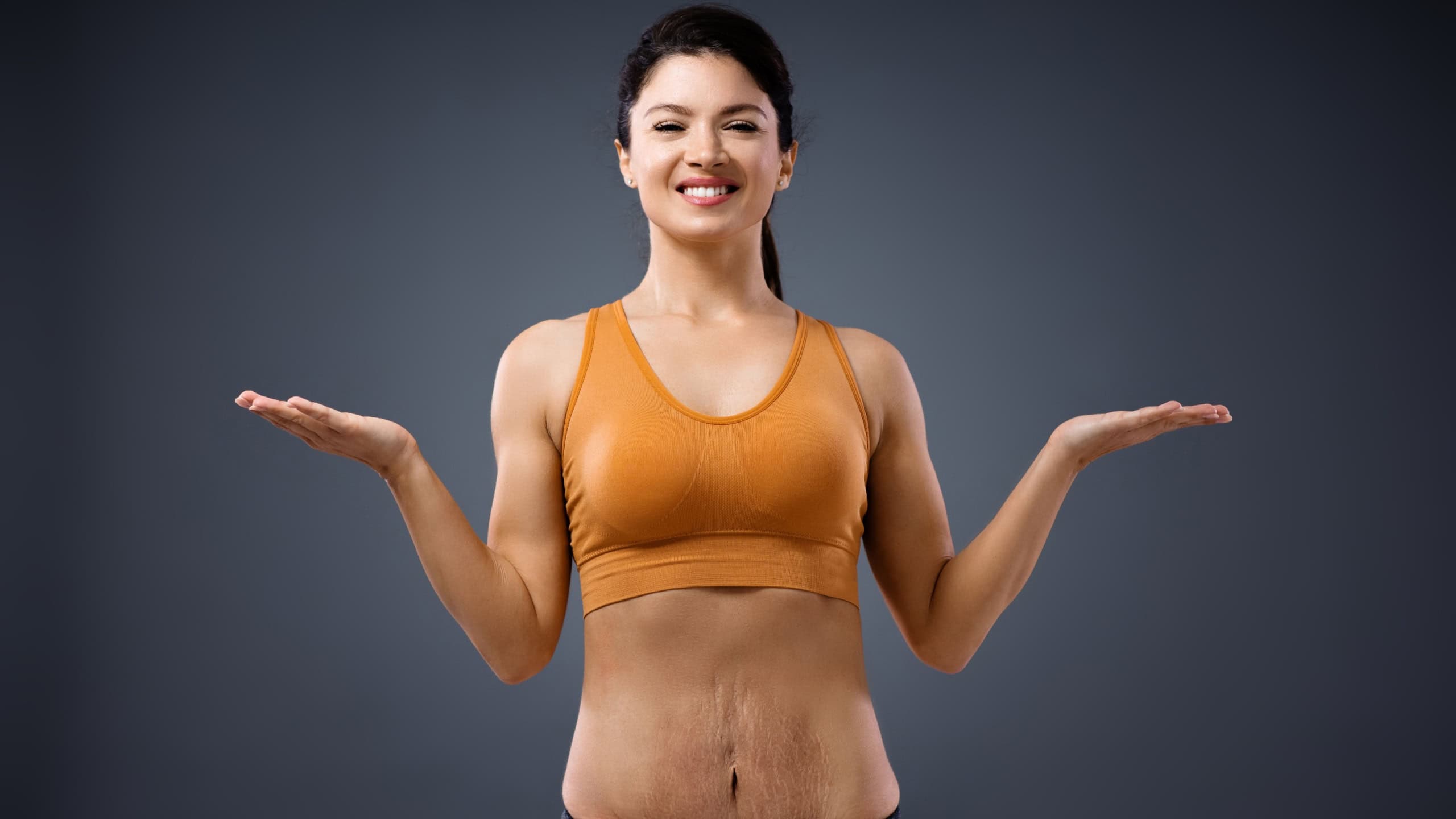 Young smiling mum standing with arms opened. She is having stretch marks on tummy and she is not having any frustrations about it.