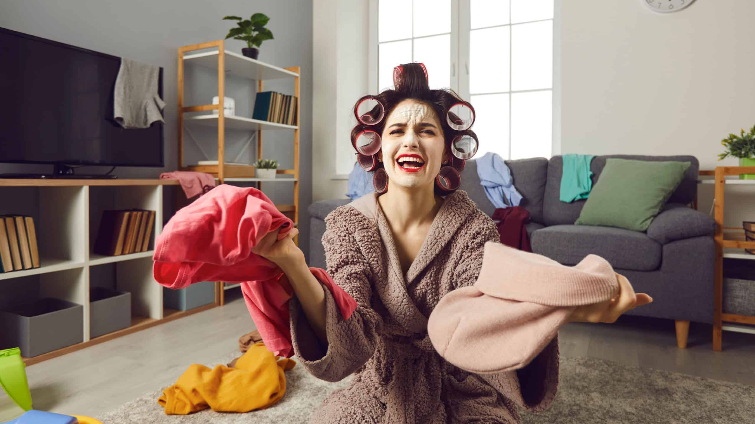 Tired desperate busy housewife cleaning up messy house. Unhappy young woman in hair curlers and skincare face mask picking piles of clothes and complaining about mess and chaos. Women's chores concept