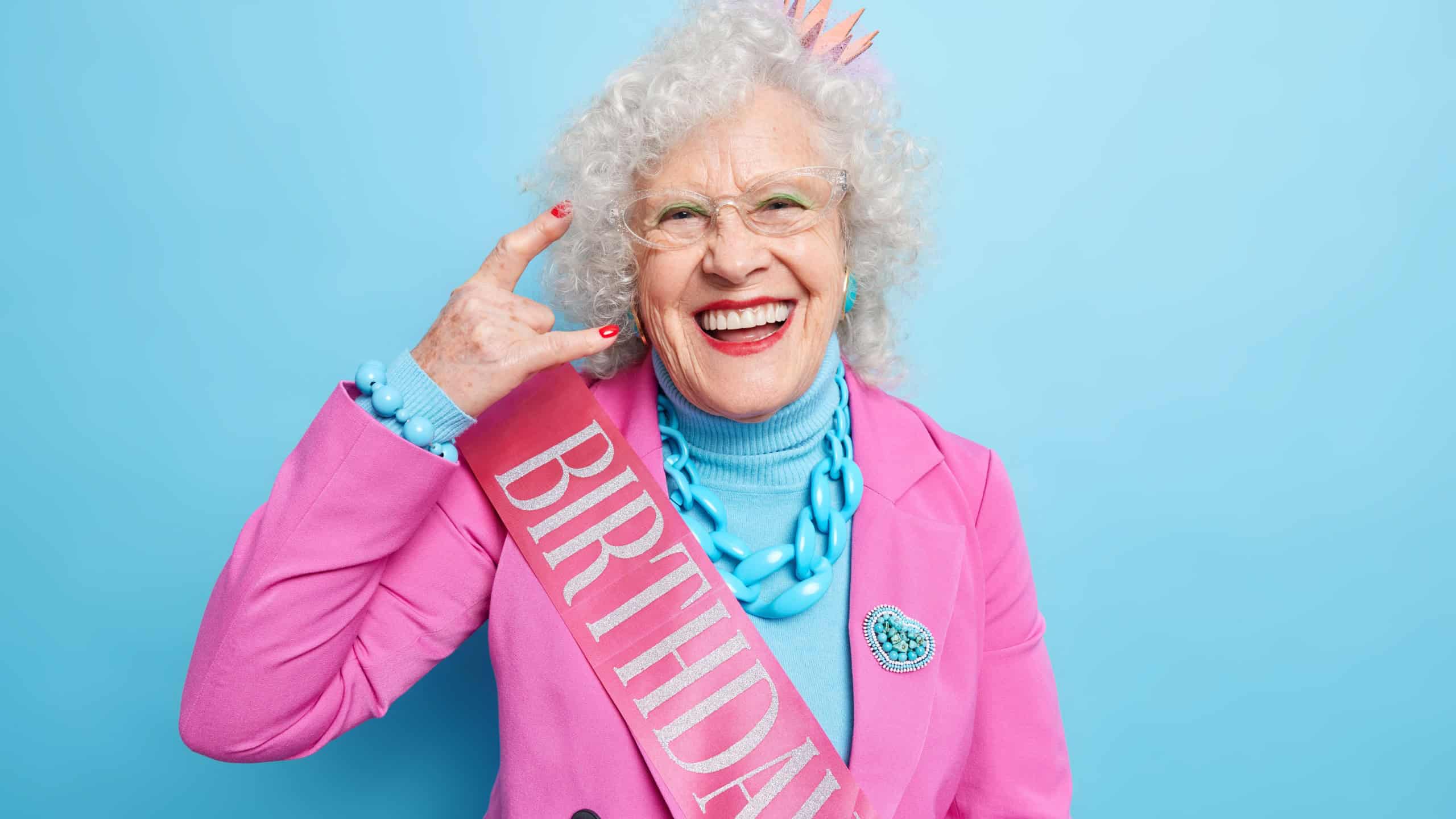 Awesome happy grey haired woman makes rock n roll gesture enjoys cool birthday party dressed in festive clothes smiles gladfully feels young again isolated over blue background. Senior rocker